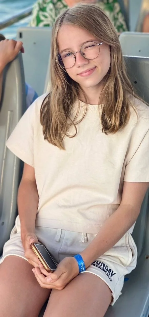 Yuliana sitting on a molded plastic bench seat outdoors, wearing round glasses and a plain cream T-shirt, her long fair hair loose, smiling with one eye closed and a phone in her hands