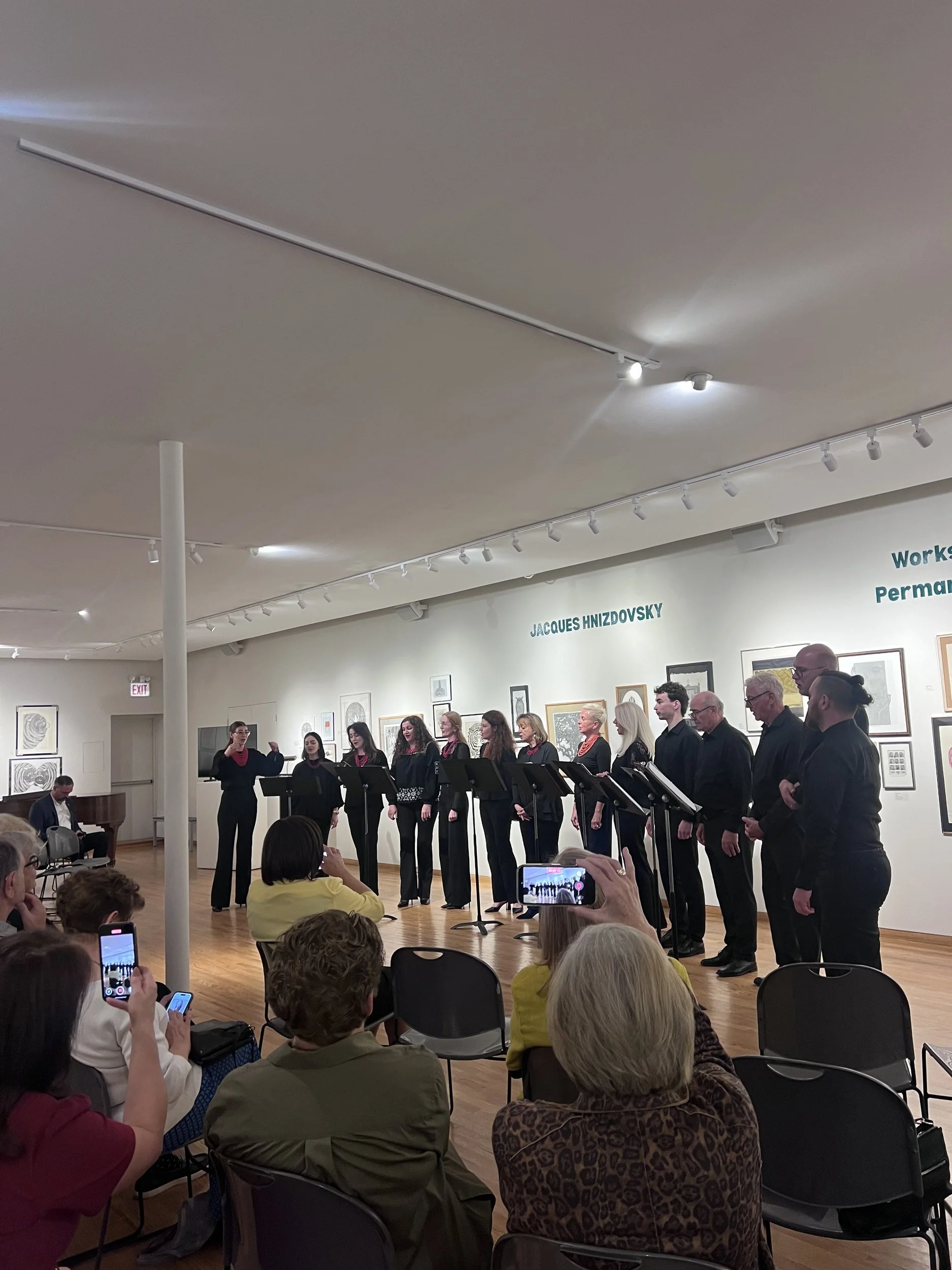Choir Glorious singers stand behind music stands as their conductor leads them before a seated audience.