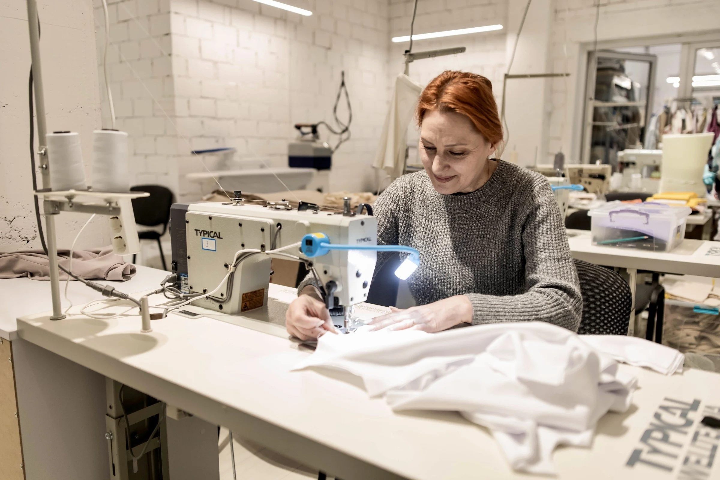 A woman in a grey jumper sits at an industrial sewing machine under a work lamp, guiding white fabric through it, in a workshop lined with more machines