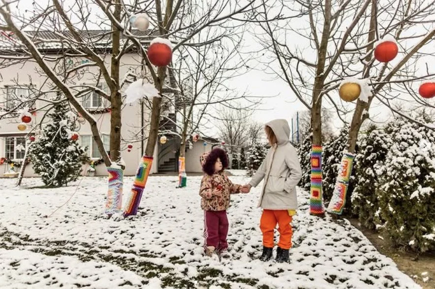 A small child and a taller person in winter coats holding hands in a snowy garden hung with knitted tree wraps and baubles