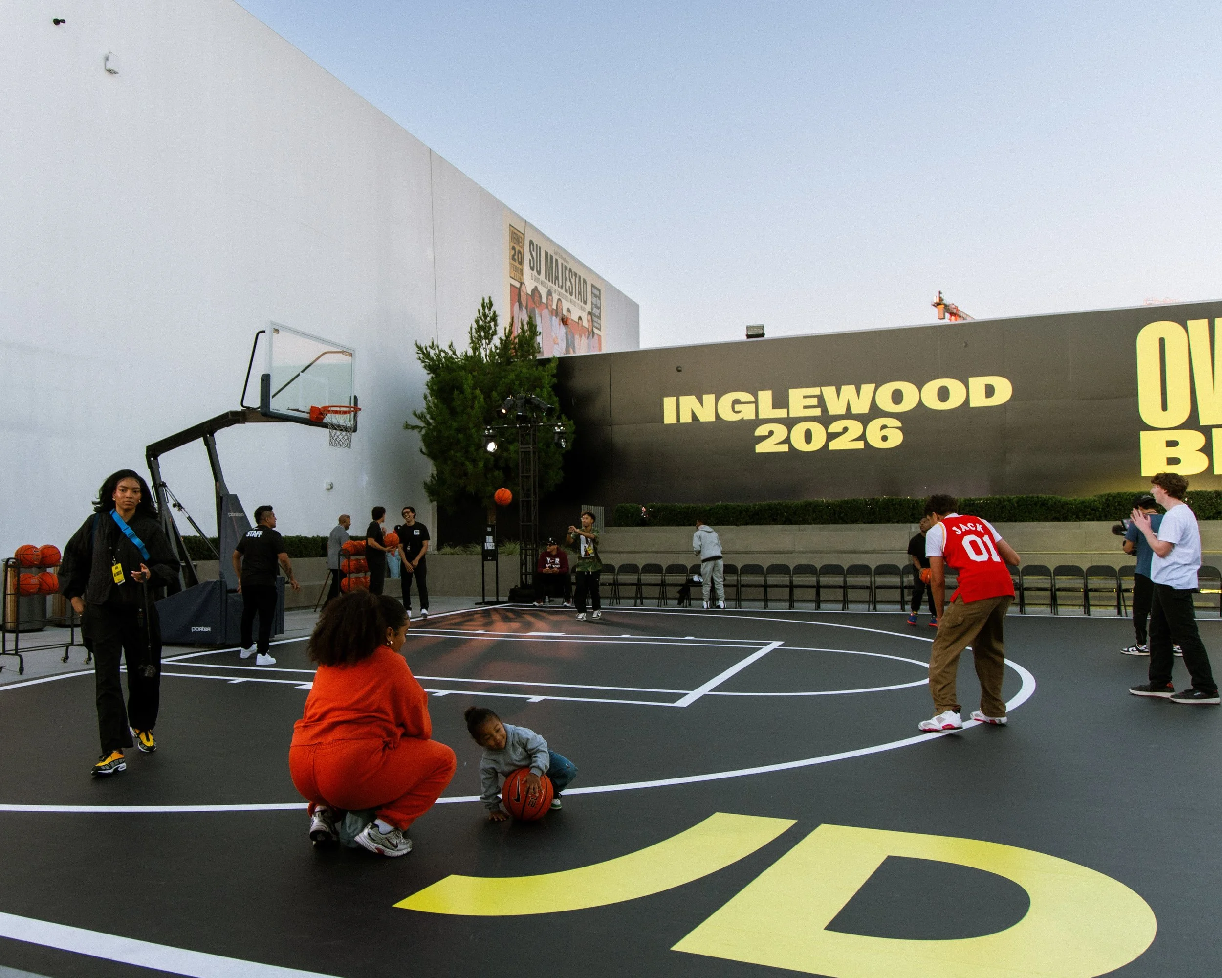 People playing basketball on an outdoor court with a large black wall displaying yellow text 'INGLEWOOD 2026' and smaller text 'OUR B' in the background.