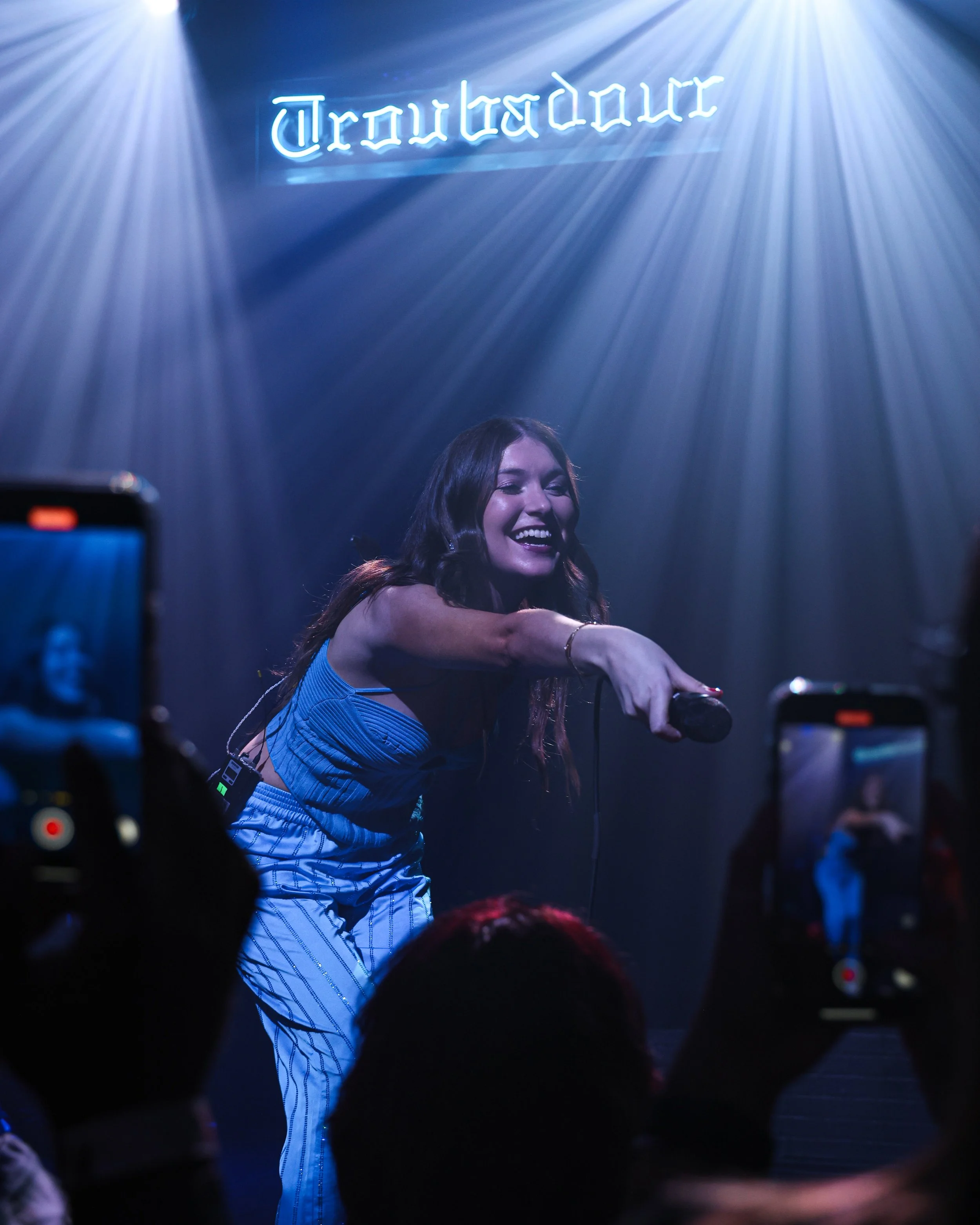A woman on stage holding a microphone, smiling, with a blue neon sign that says "Troubadour" in the background, surrounded by audience members taking photos.