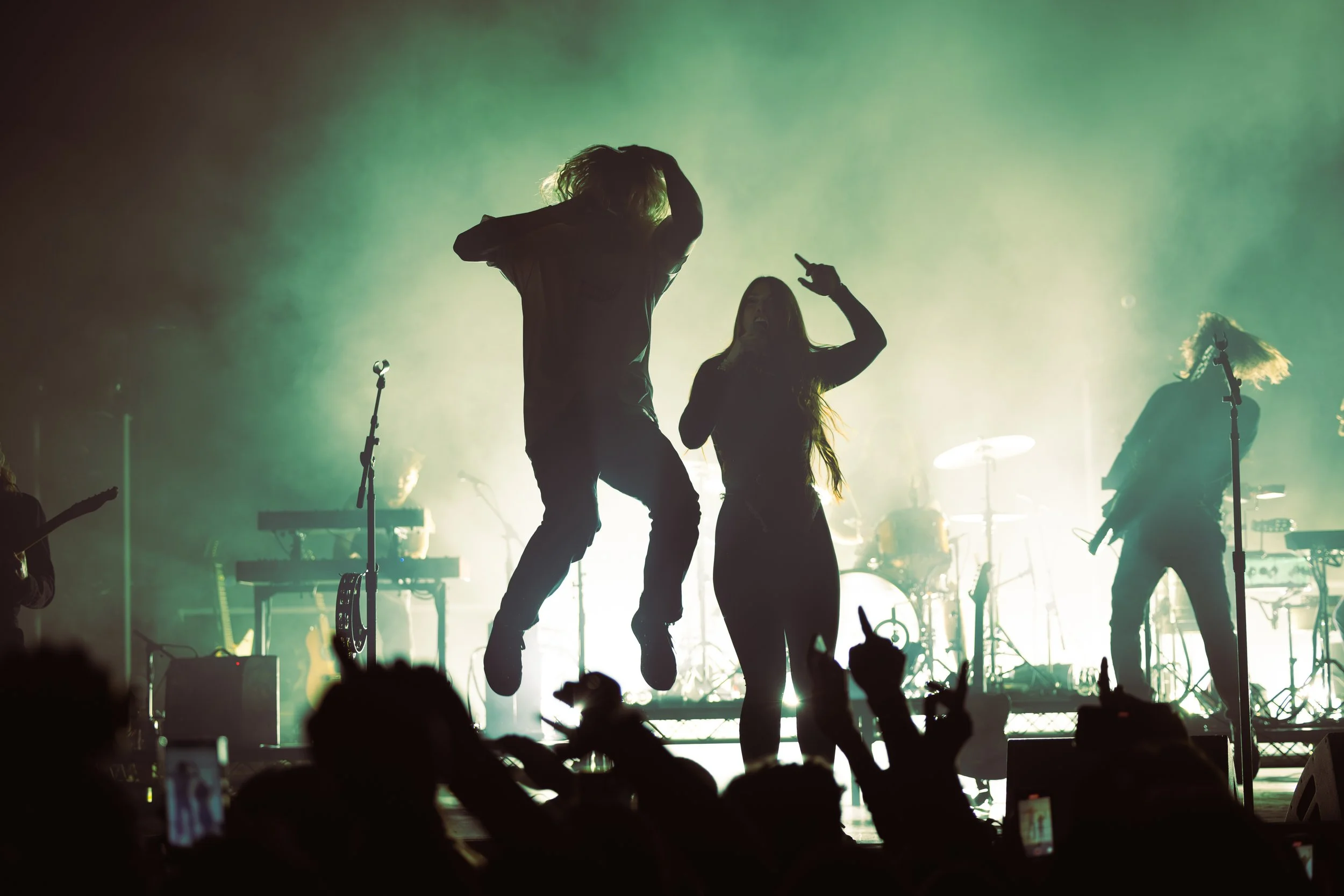 Silhouettes of performers dancing and singing on stage during a concert with green backlighting and musical instruments in the background.