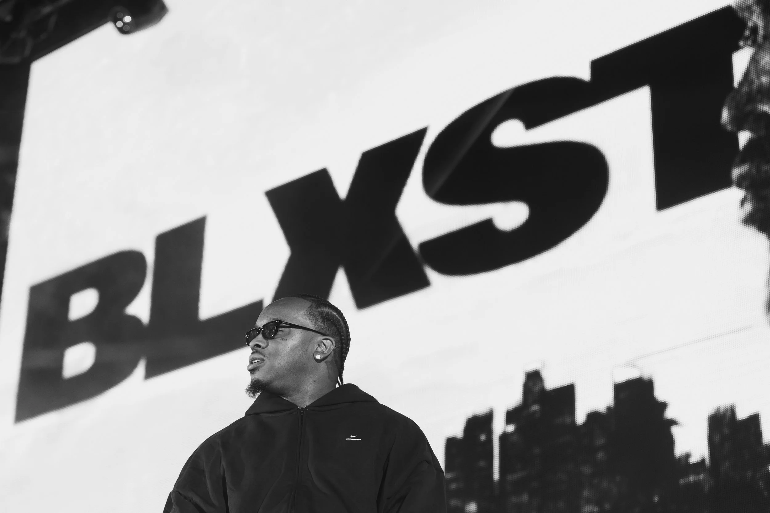 A man wearing sunglasses and a black Nike hoodie stands in front of a large screen displaying the Blxst logo with a city skyline silhouette in the background, in black and white.