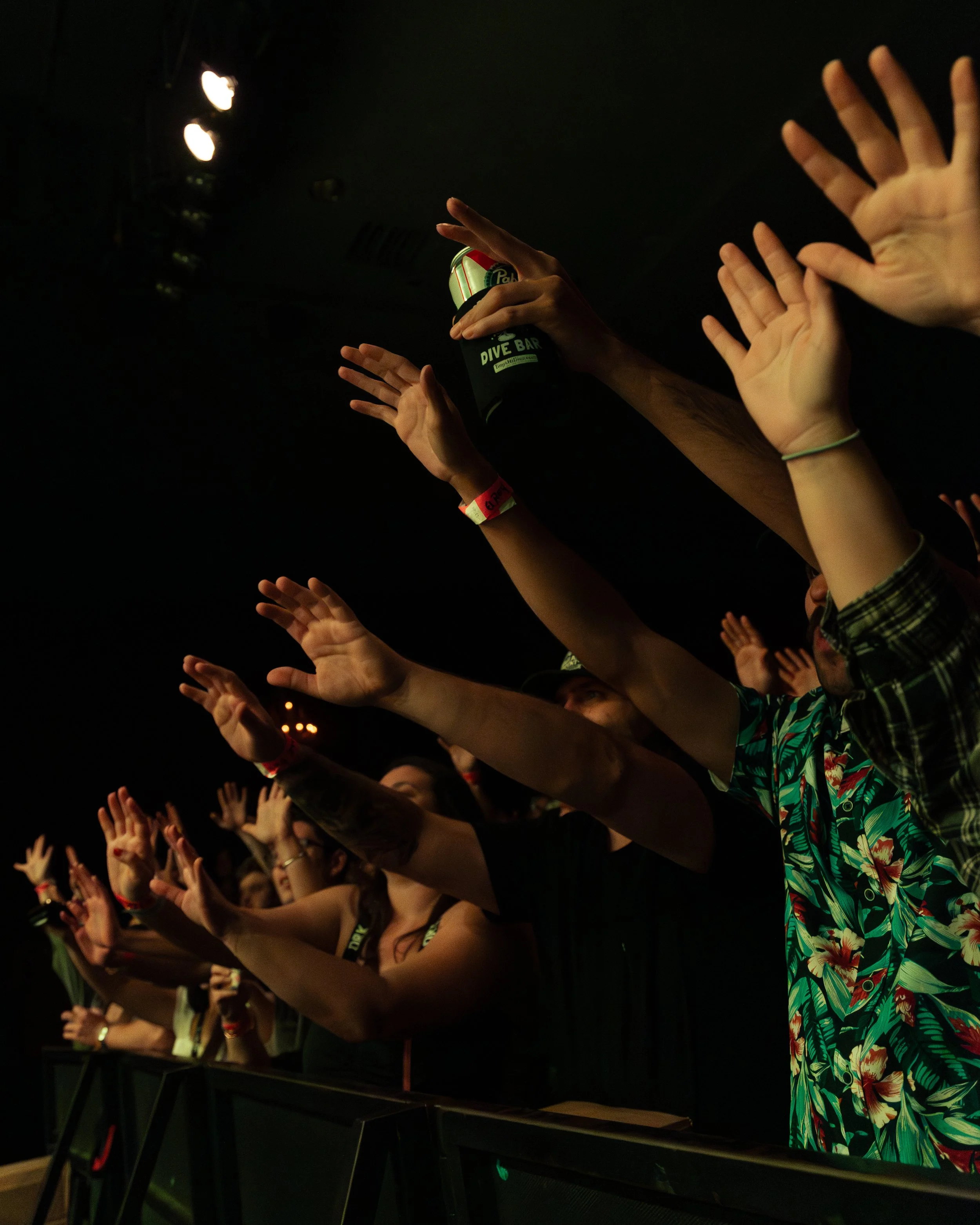 People raising their hands at a concert or event, with some holding drinks, in a dark indoor setting.