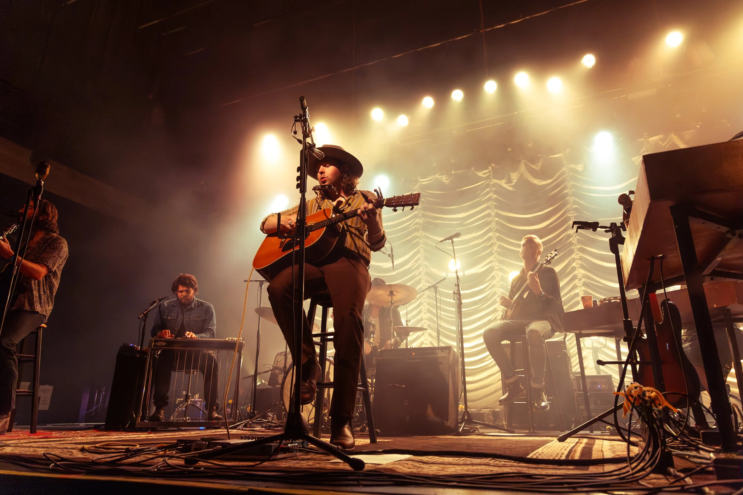 Musicians performing on stage with guitars in front of a textured curtain with bright stage lights behind them.