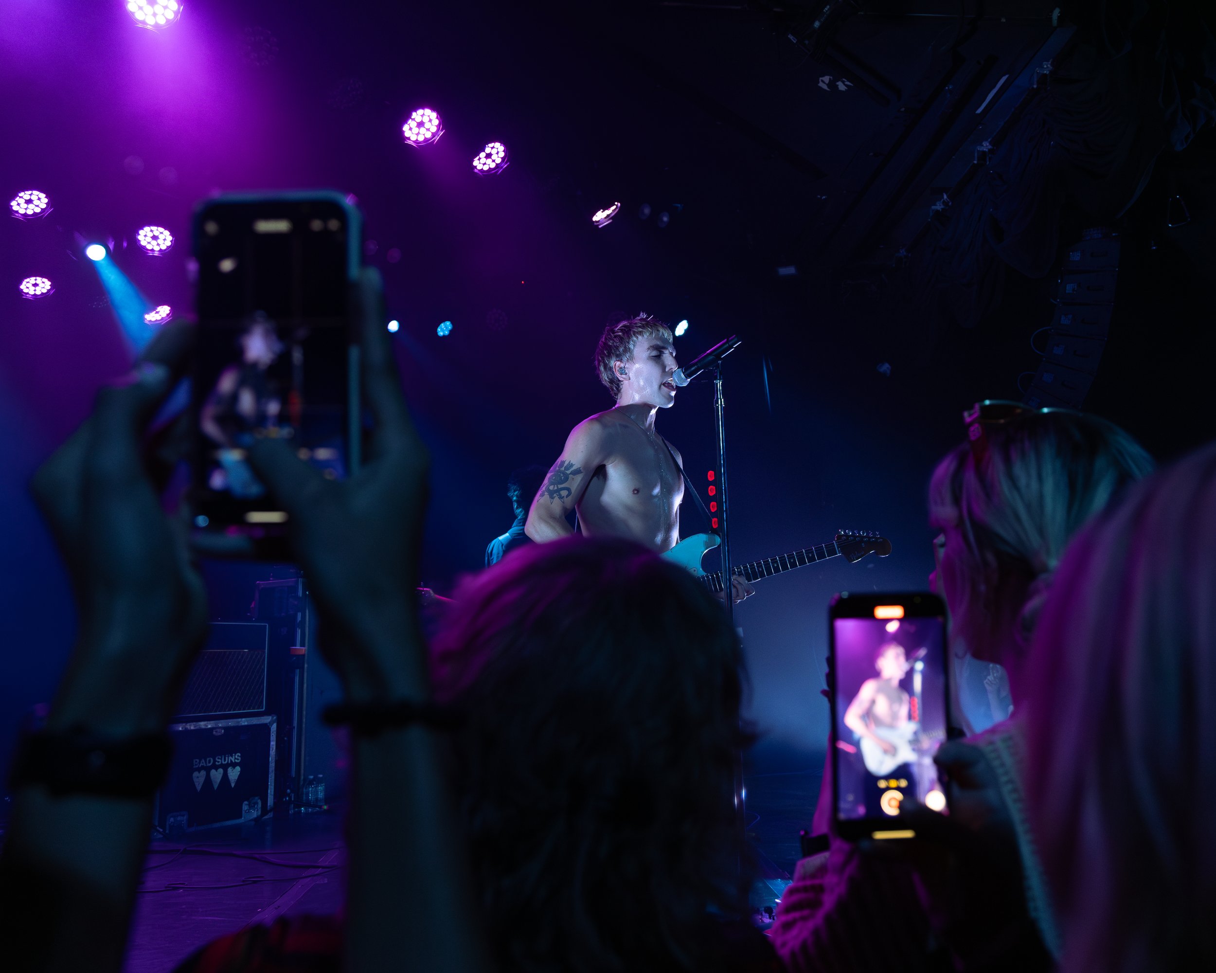 A shirtless male musician with tattoos on his arm singing into a microphone while playing an electric guitar on stage with purple and blue lighting, audience members are taking photos and videos.