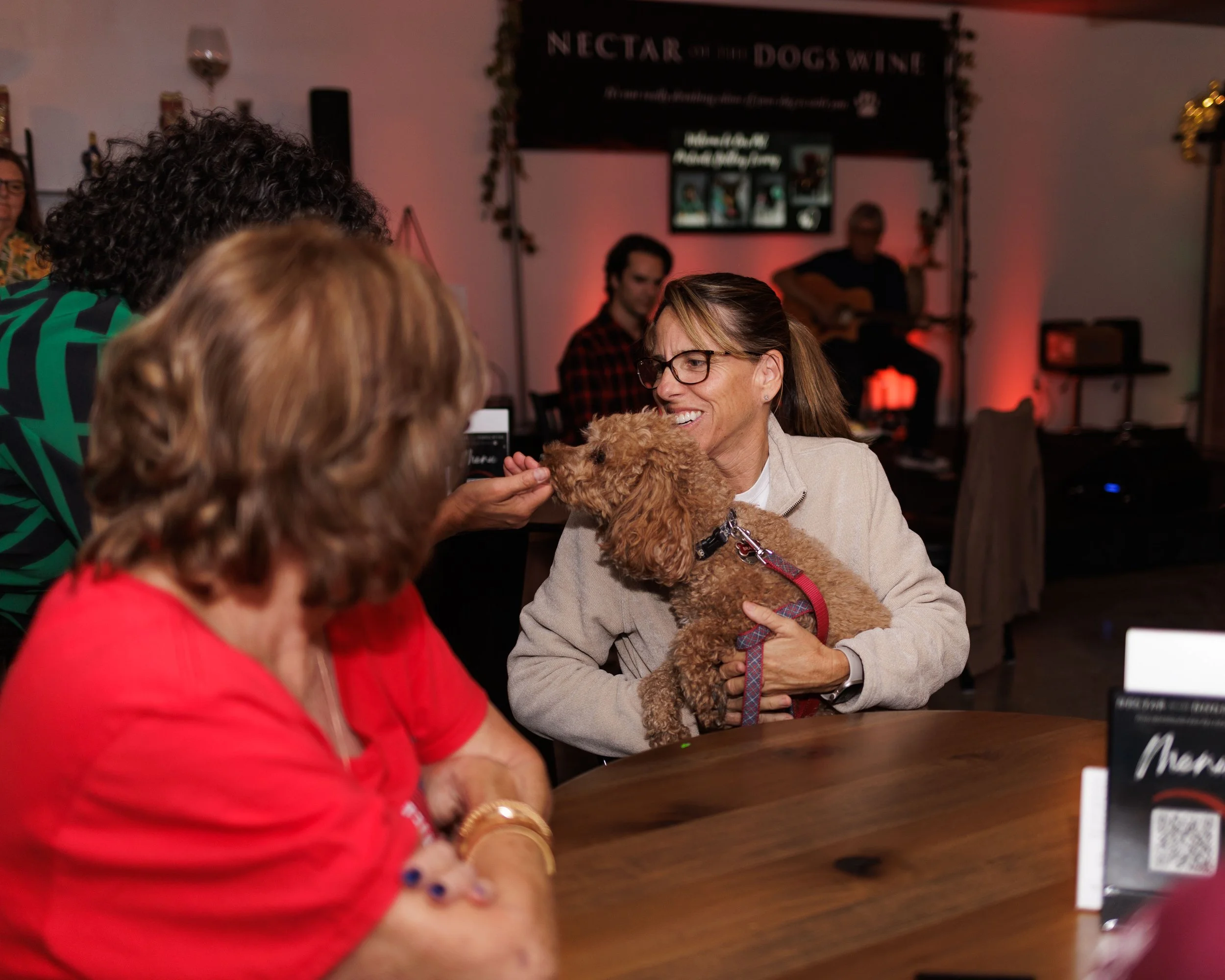 Two women, one with red hair wearing a red shirt and the other with glasses and a beige jacket, are smiling and interacting with a small brown poodle dog at a social gathering, with a band playing live music in the background.