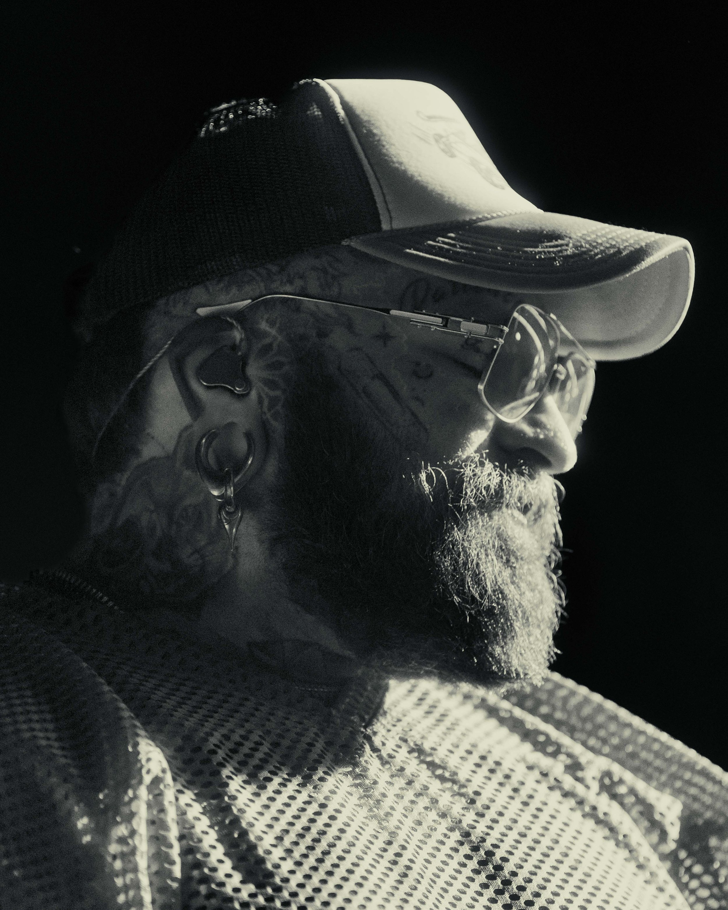 A man with sunglasses and a cap looking to the right, featuring tattoos, piercings, and a beard, in a black and white photo.