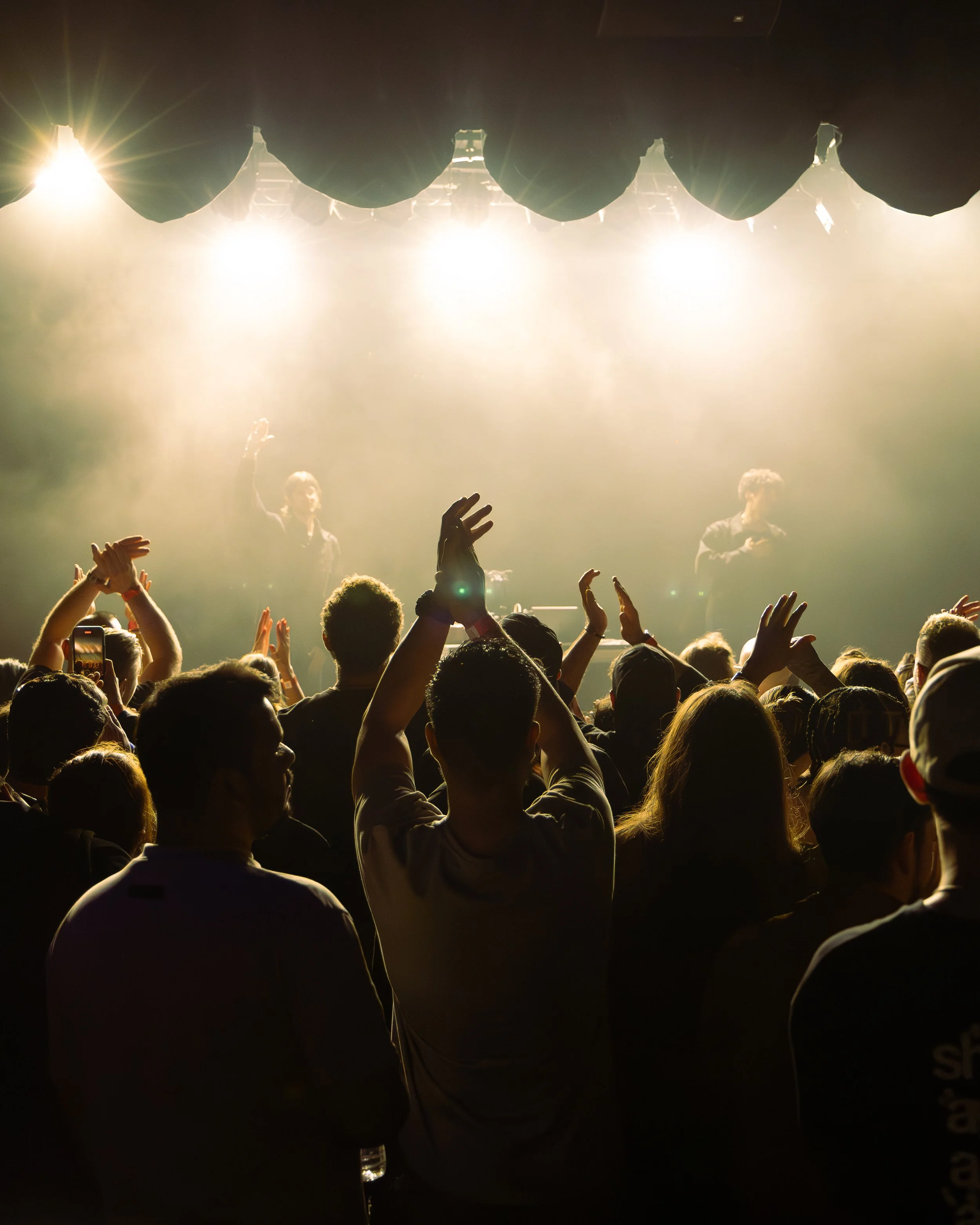 A crowd of people at a concert or performance, with bright stage lights shining and two performers visible on stage in the background.