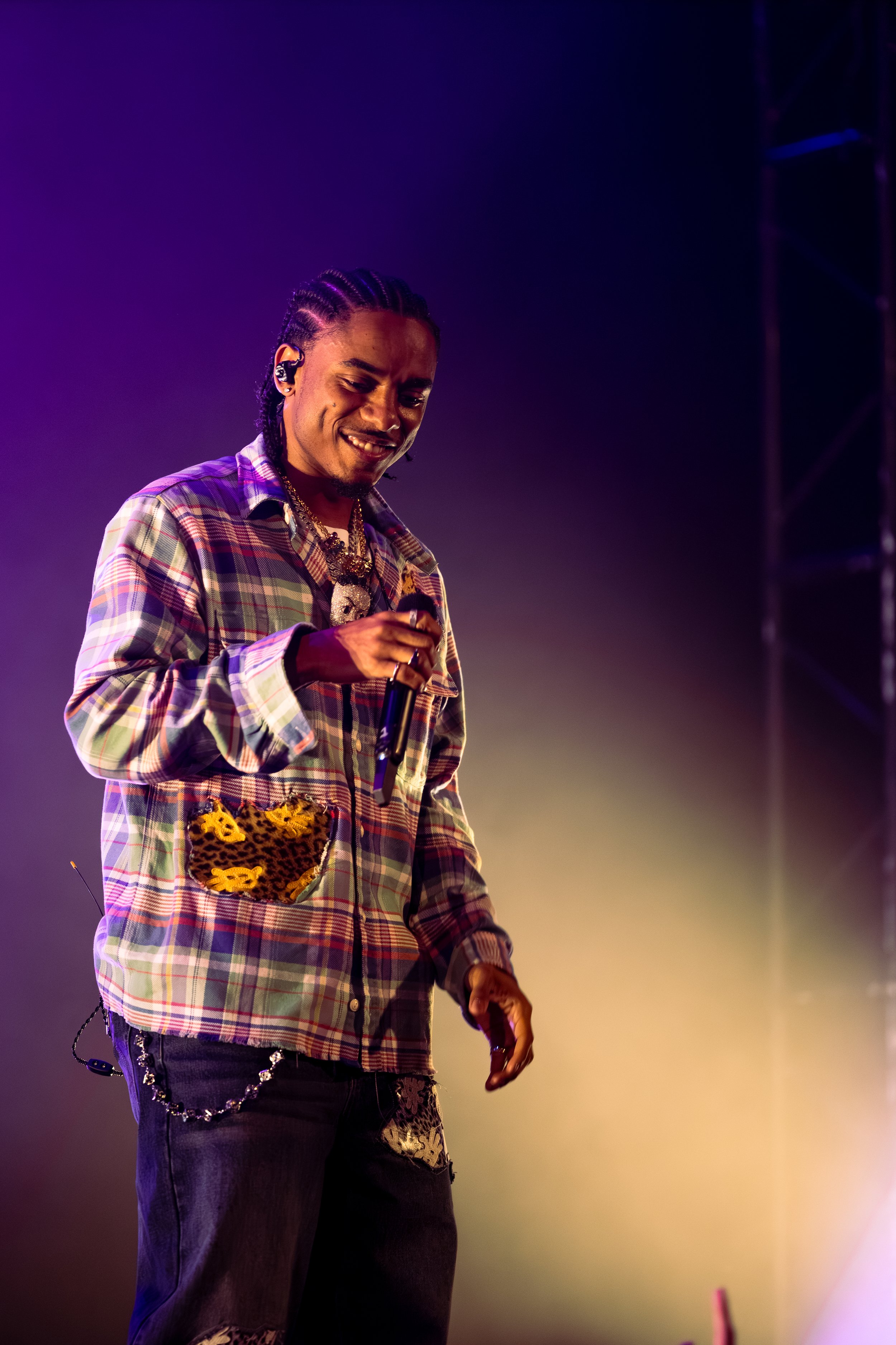 Man with braided hair wearing a plaid jacket with leopard print pocket holding a microphone on stage with purple and yellow lighting