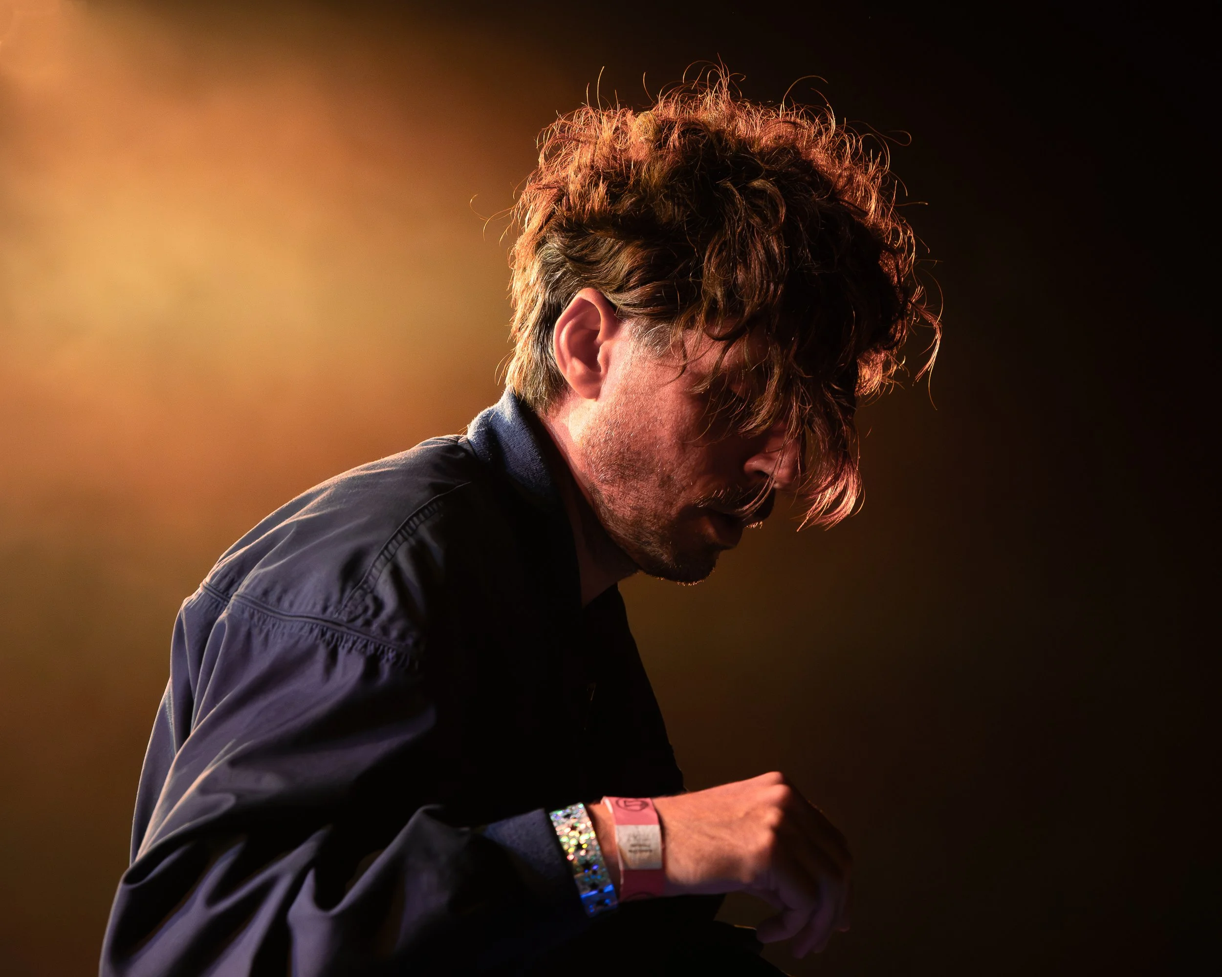 A man with messy hair and glasses, wearing a black jacket, looking down, illuminated by warm stage lighting.