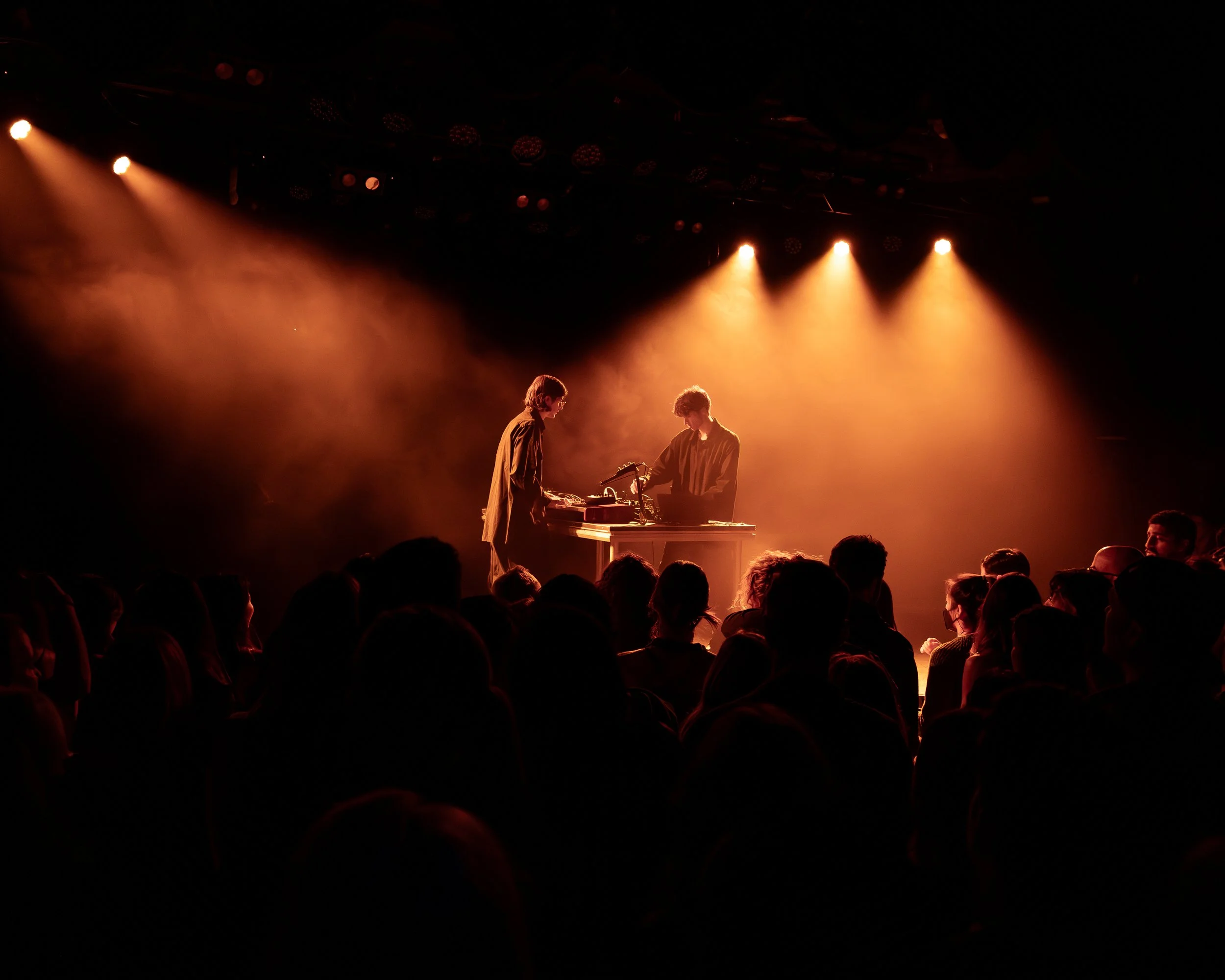 Two musicians performing on stage with orange stage lights and an audience watching.