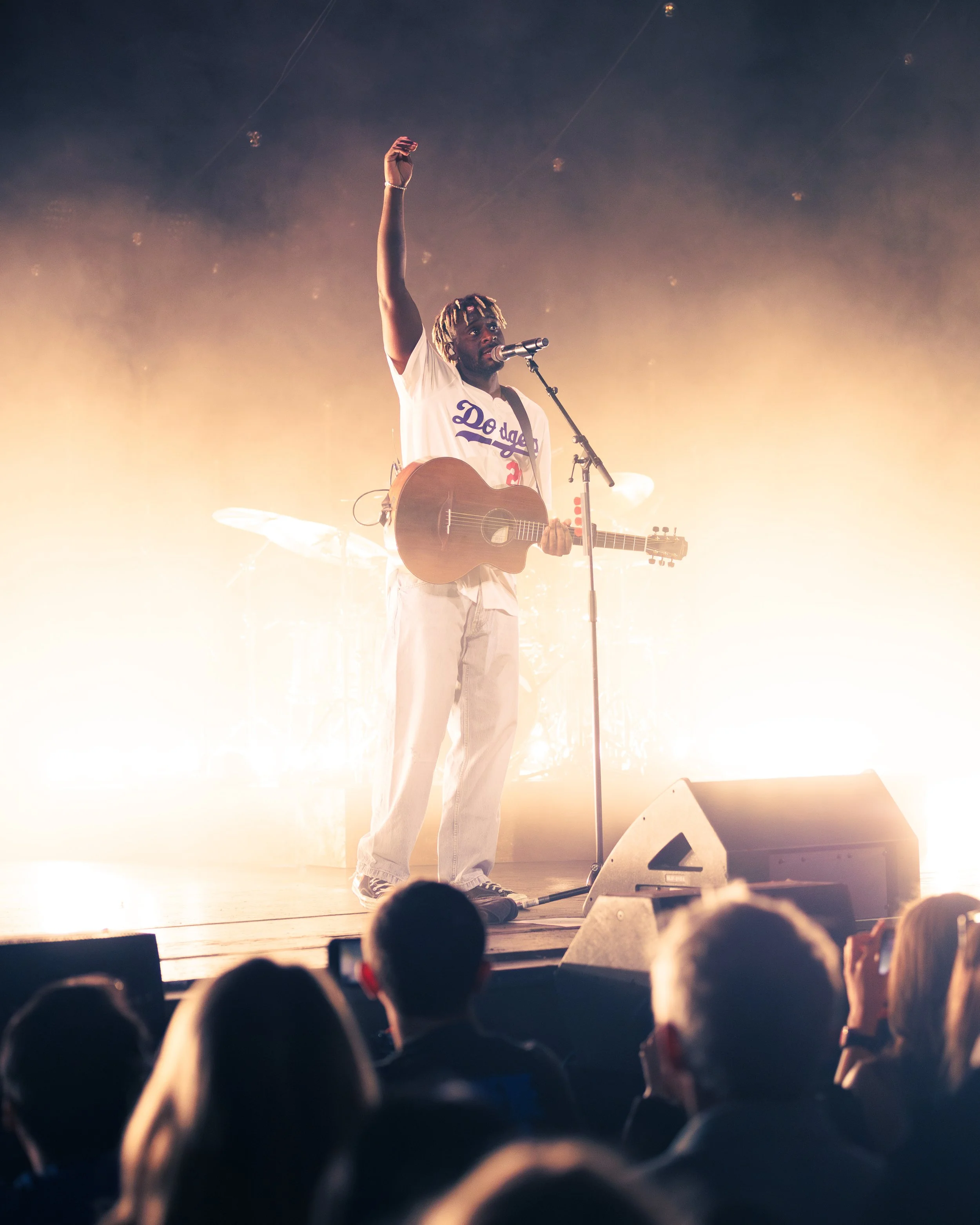 A male performer with dreadlocks on stage singing into a microphone and playing an acoustic guitar, with bright stage lights behind him, in front of an audience.