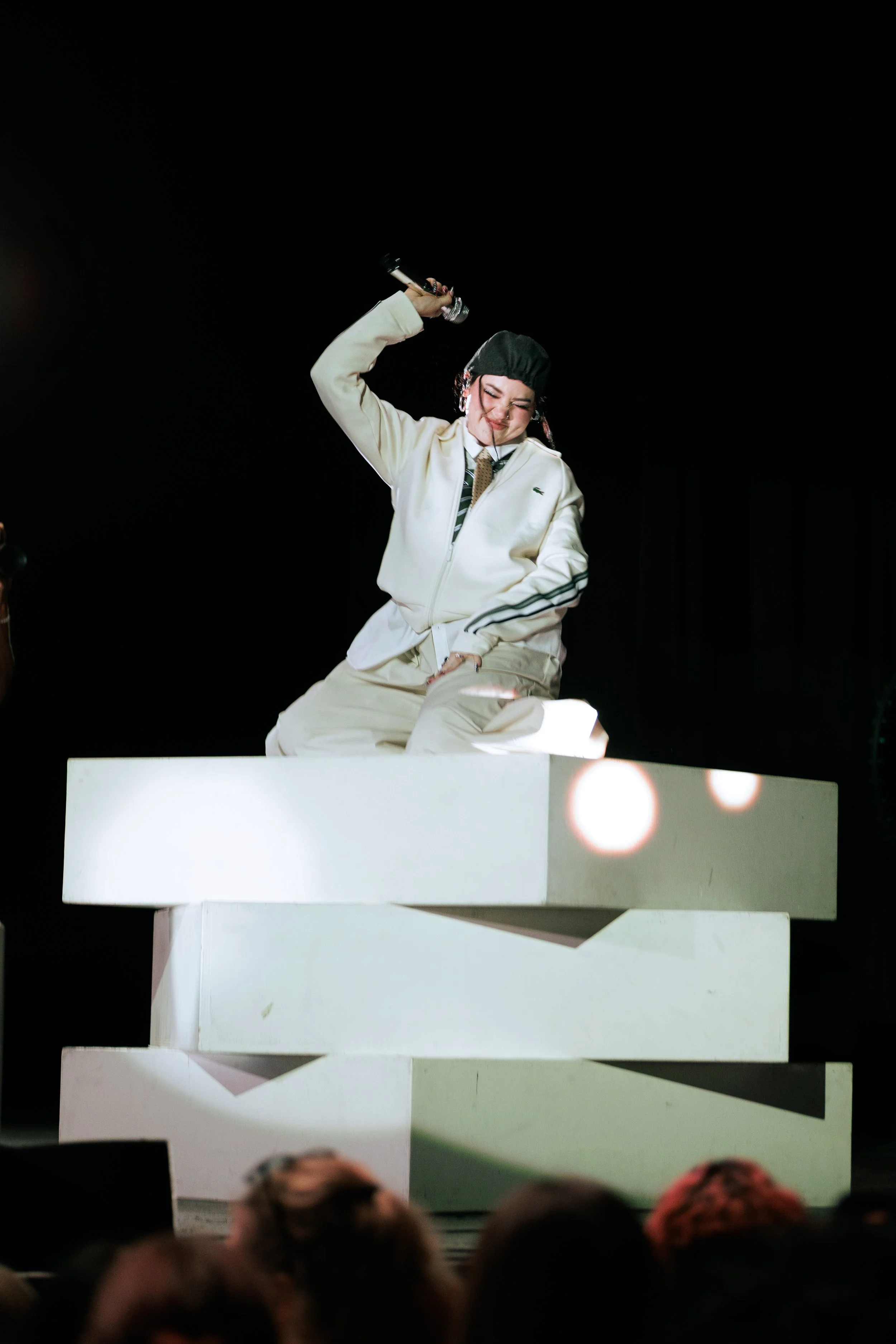 A woman performing on stage, kneeling on a white platform, holding a microphone in her right hand, with a big smile, wearing a white jacket, beige pants, black cap, and a tie, against a dark background.