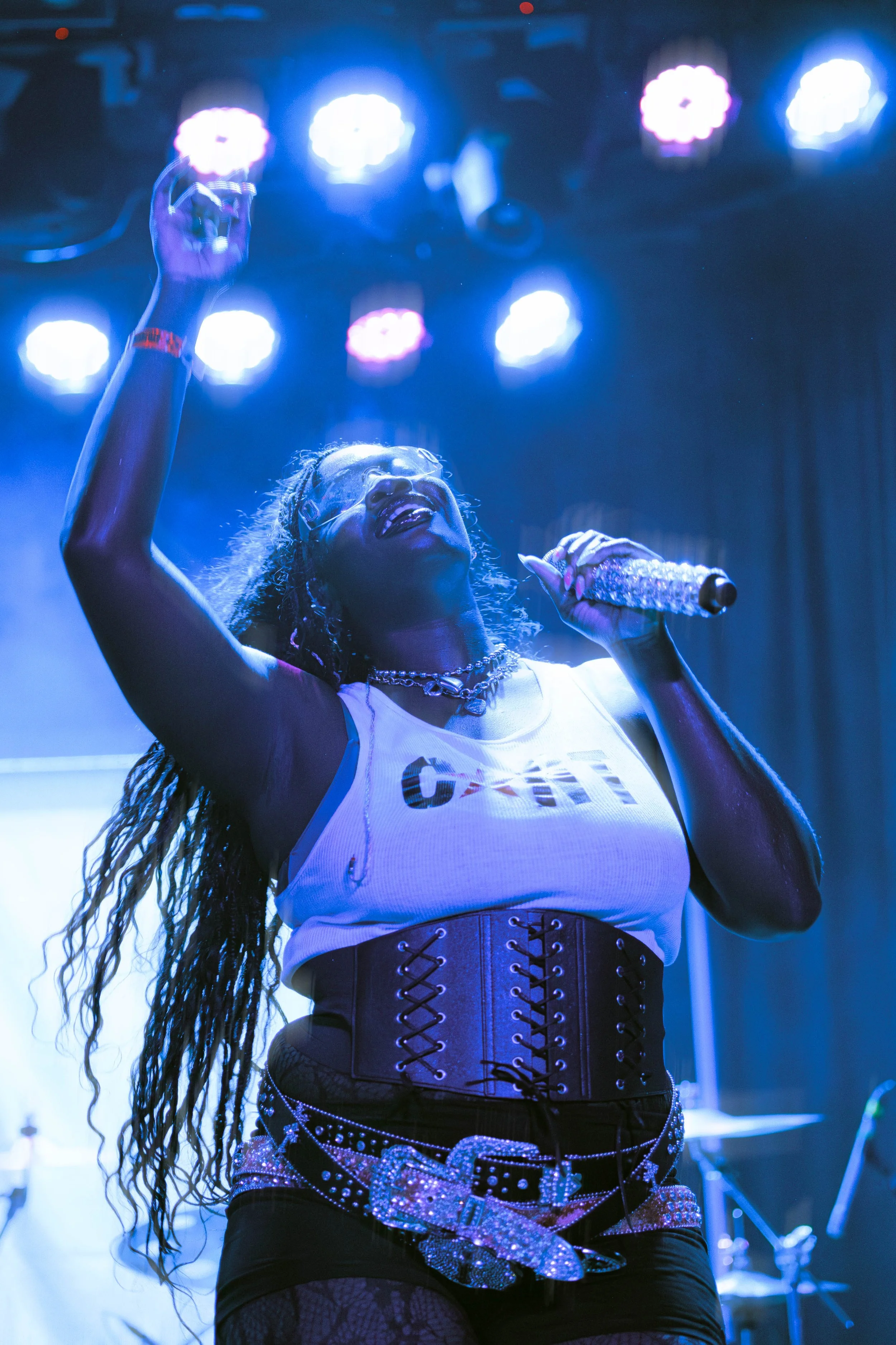Performer singing on stage with bright overhead lights, wearing a white tank top, a wide belt with a large rhinestone buckle, and fishnet stockings, holding a glittery microphone, with one arm raised.