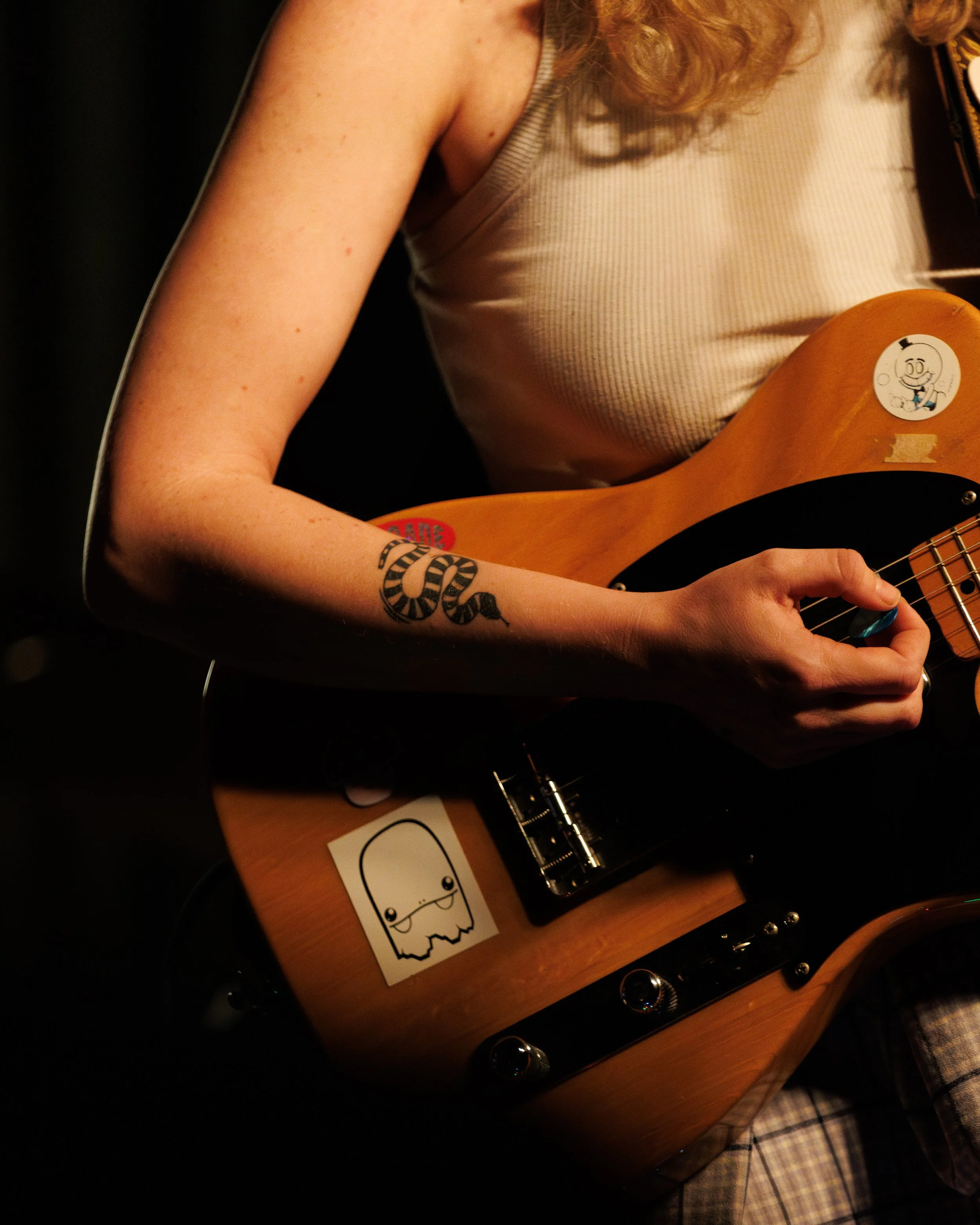 Close-up of a person with a tattoo on their arm, playing an electric guitar with stickers on it, wearing a sleeveless shirt.