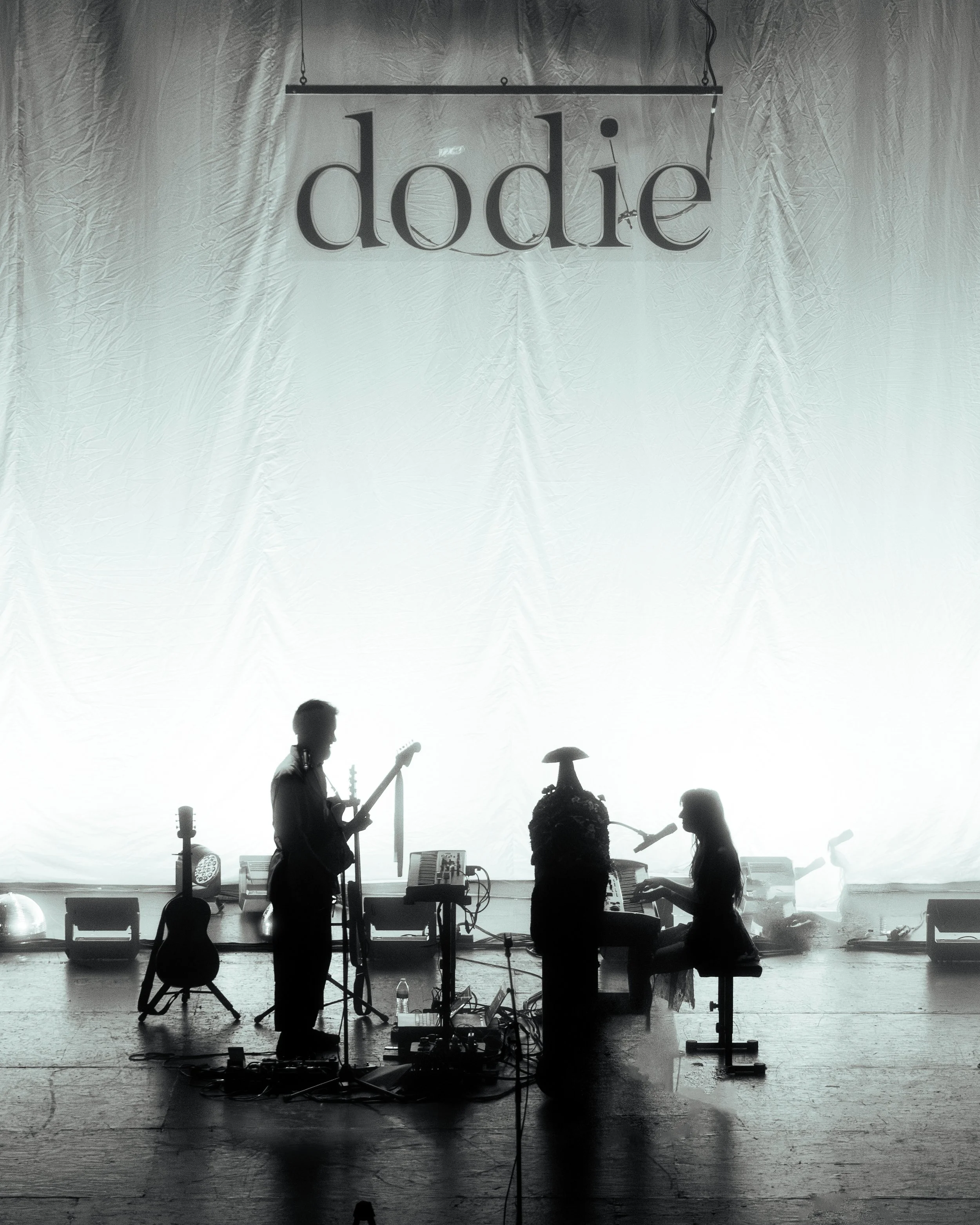Silhouettes of two musicians performing on stage with musical instruments against a bright white backdrop, with a large hanging sign that reads 'dodie' above them.