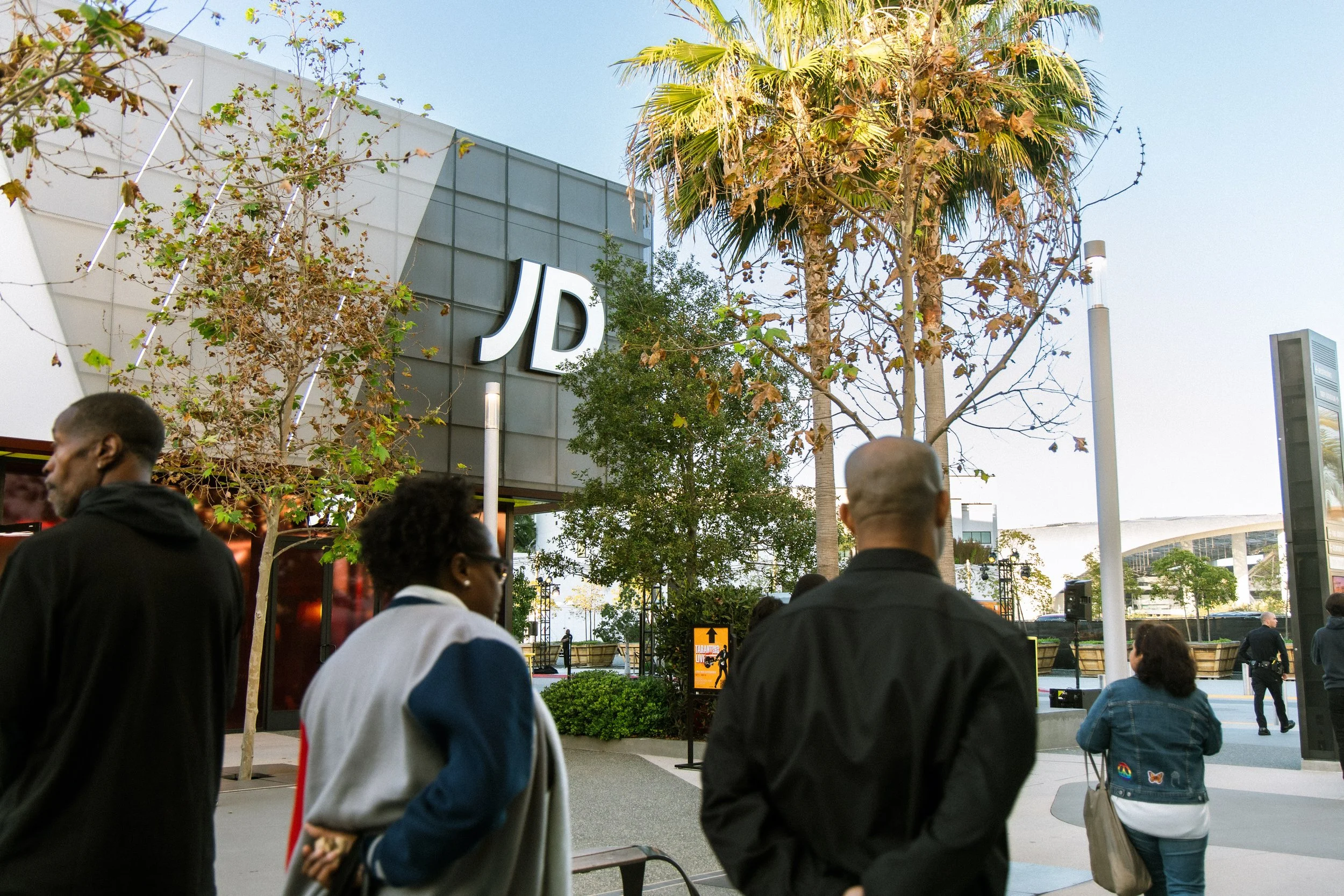People walking outside of a shopping mall with a prominent JD store sign, trees, and clear sky in the background.