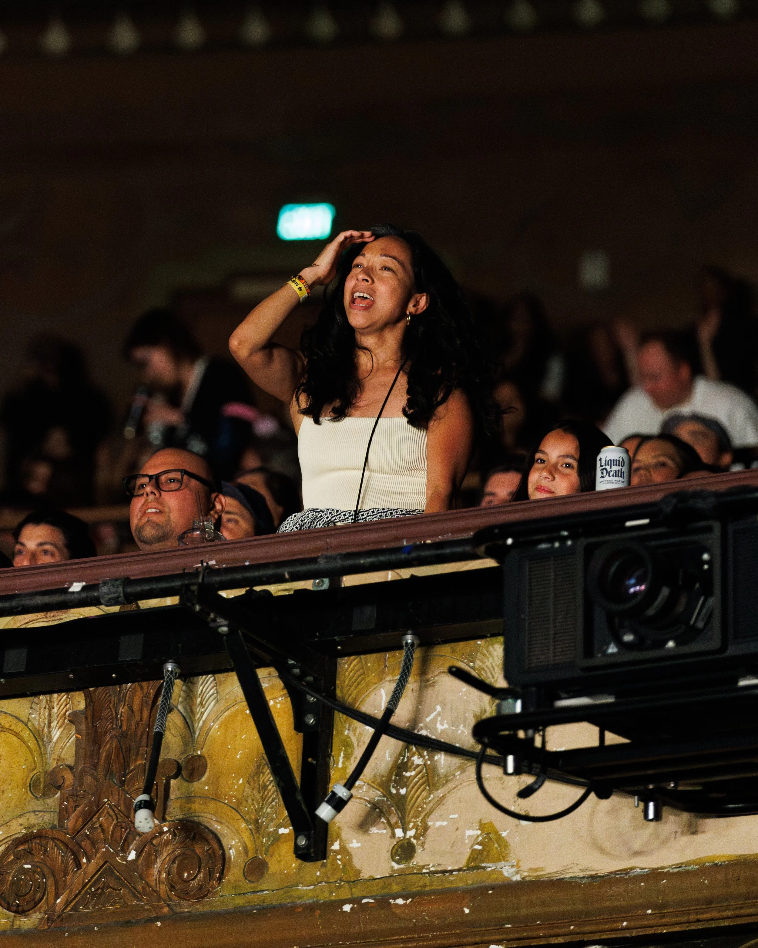 A woman in a white strapless top and patterned pants is standing with her hand on her head, appearing emotional or distressed, while attending a concert or event with a crowd around her.