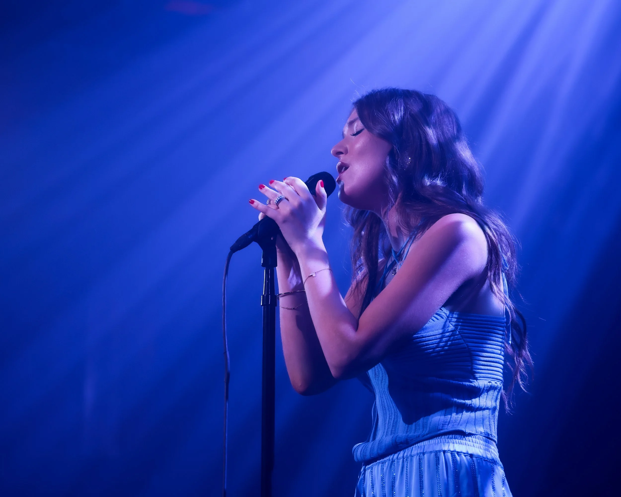 A woman singing into a microphone on stage with blue lighting and a smoky background.