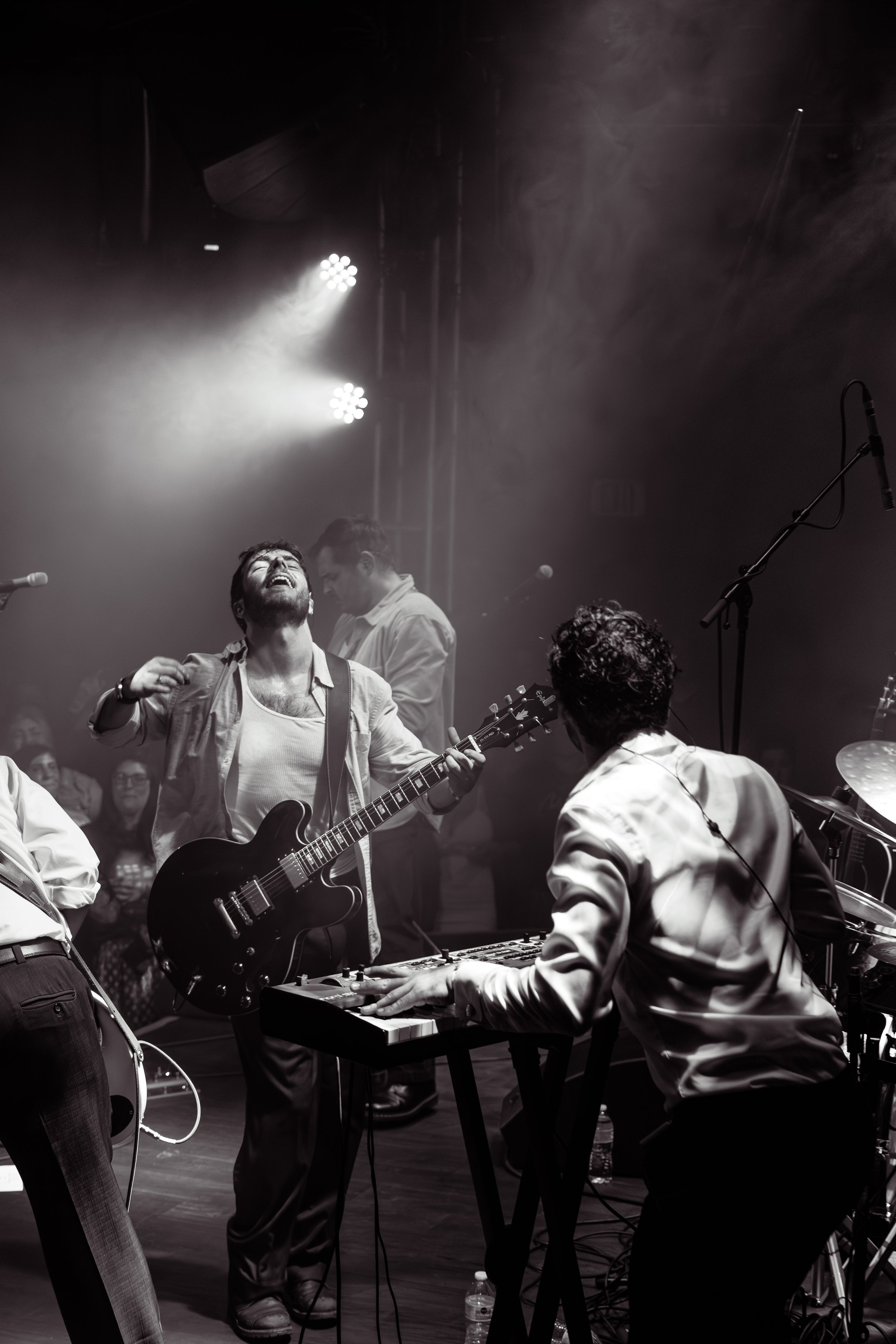 A black and white photo of a live music performance featuring a musician with a guitar singing passionately, a keyboardist playing, and a bassist in the background surrounded by an audience.