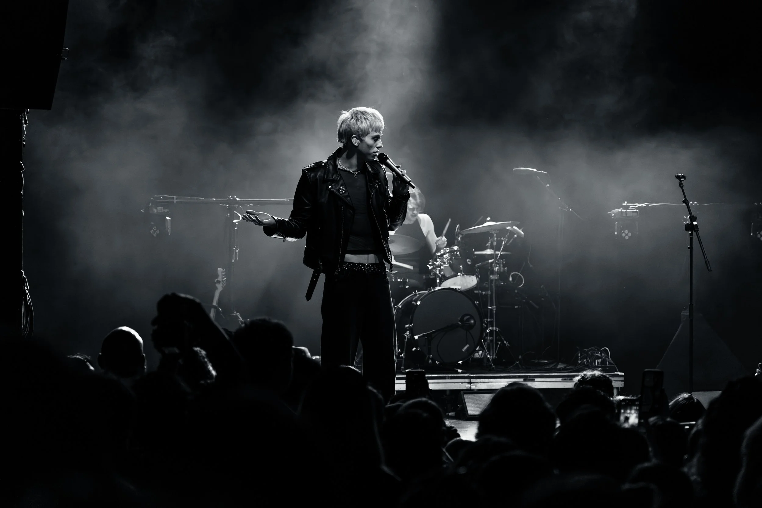 A black and white photo of a female singer on stage with a microphone, wearing a leather jacket, with her hand raised. A drummer is seen playing in the background, and there are audience members in the foreground.