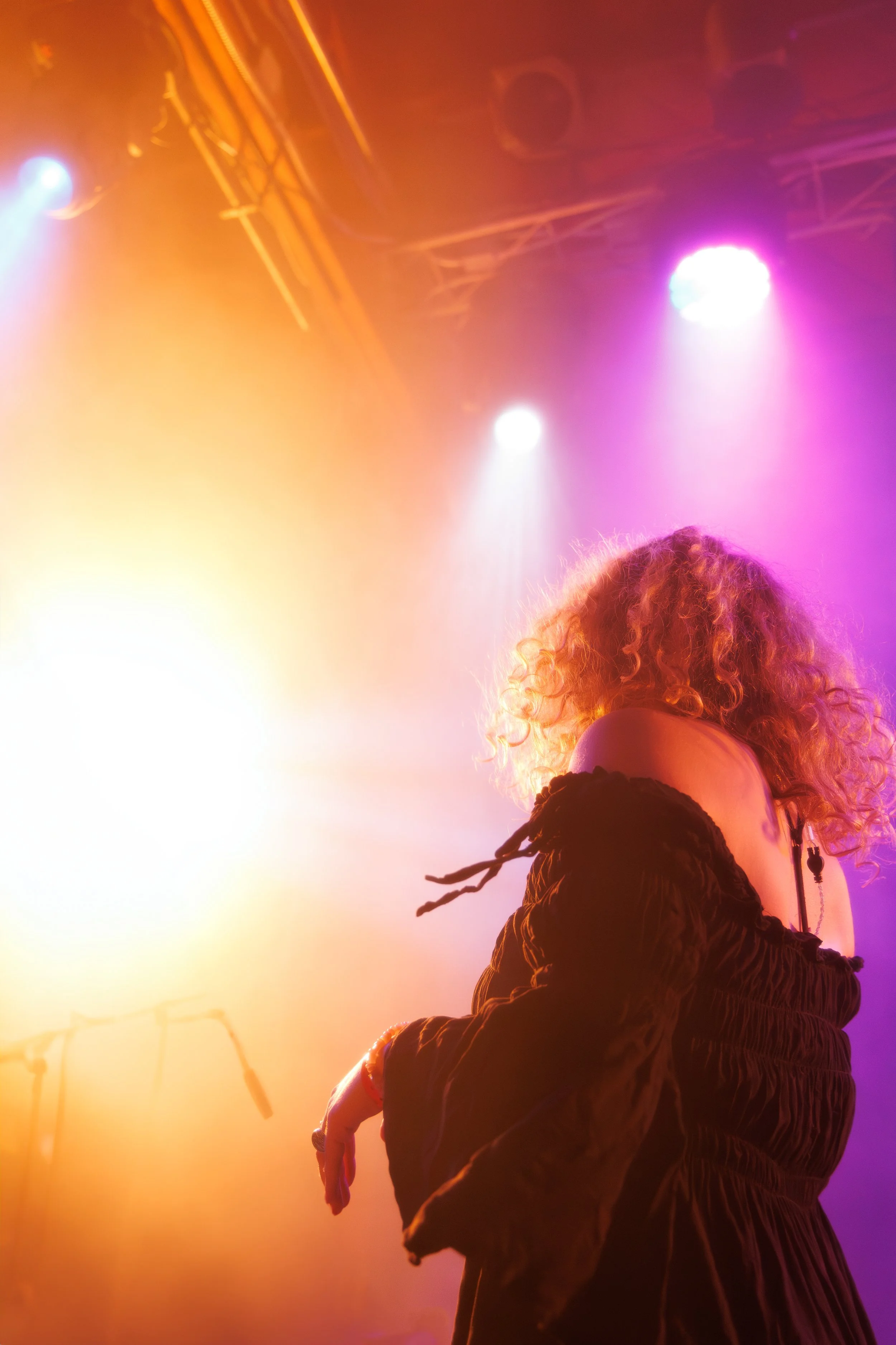 Performer with curly hair on stage illuminated with colorful pink, purple, and orange lights