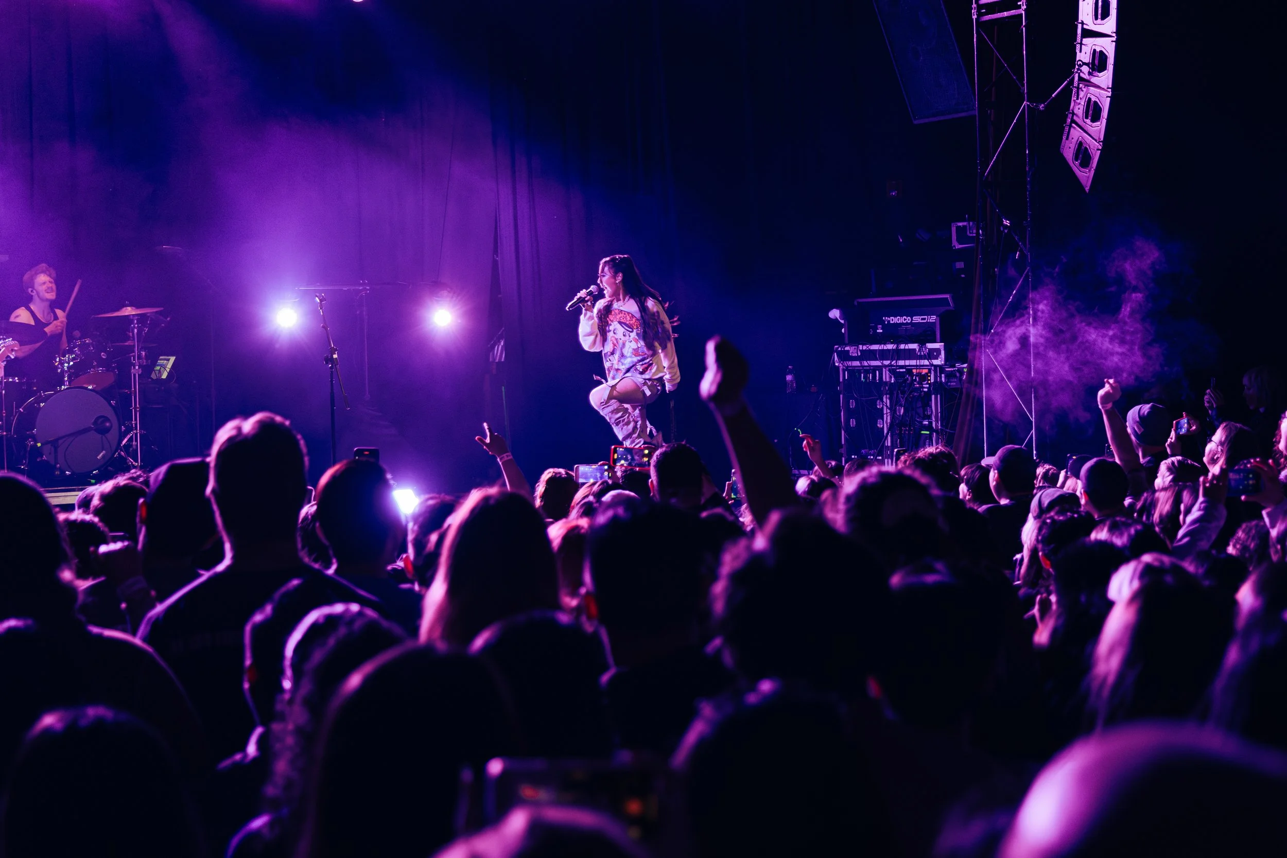 A singer performing on stage during a concert with purple lighting, with an audience watching and recording.