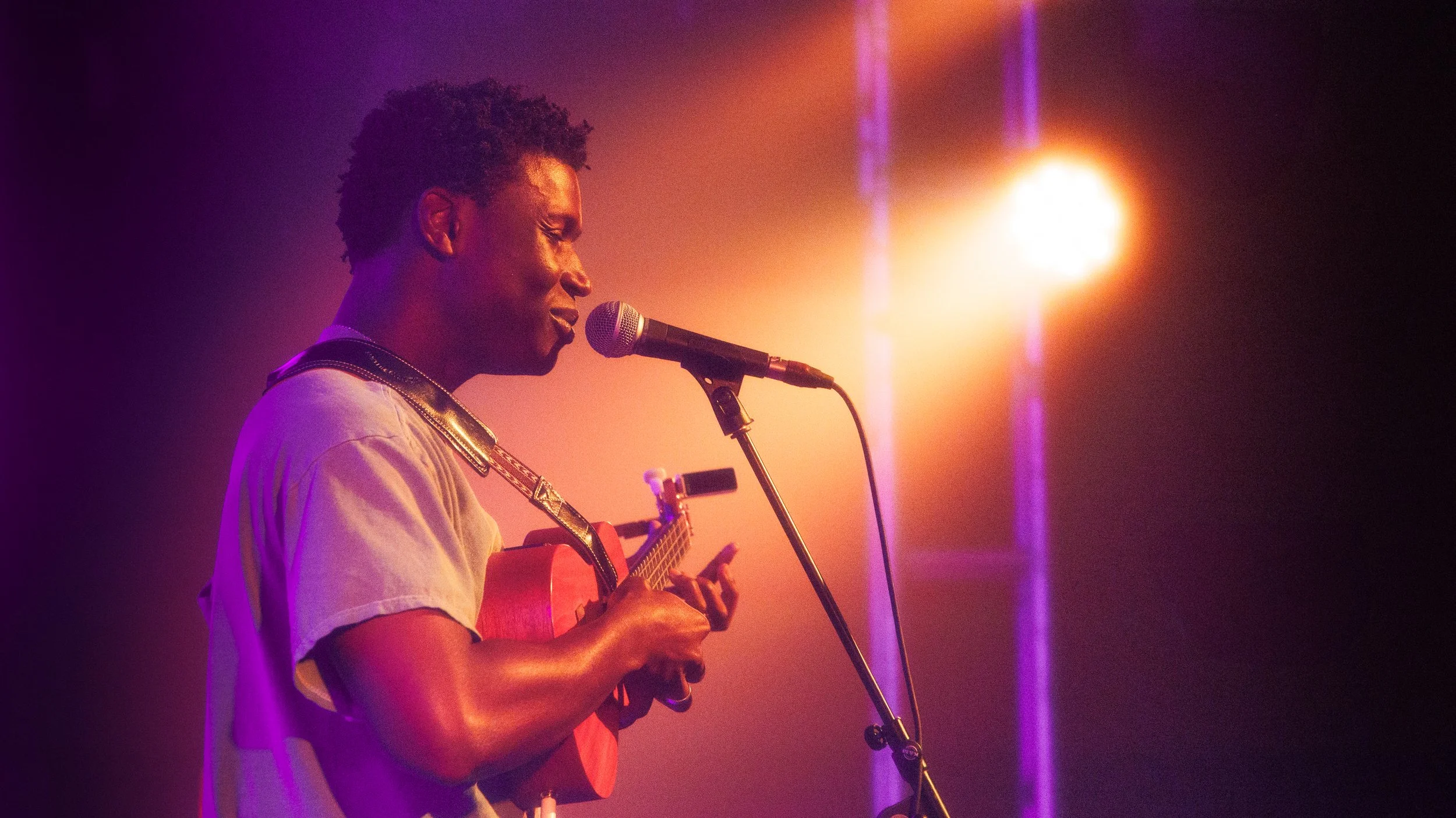 A young person with short curly hair performing on stage, playing a ukulele and singing into a microphone, under warm stage lighting.