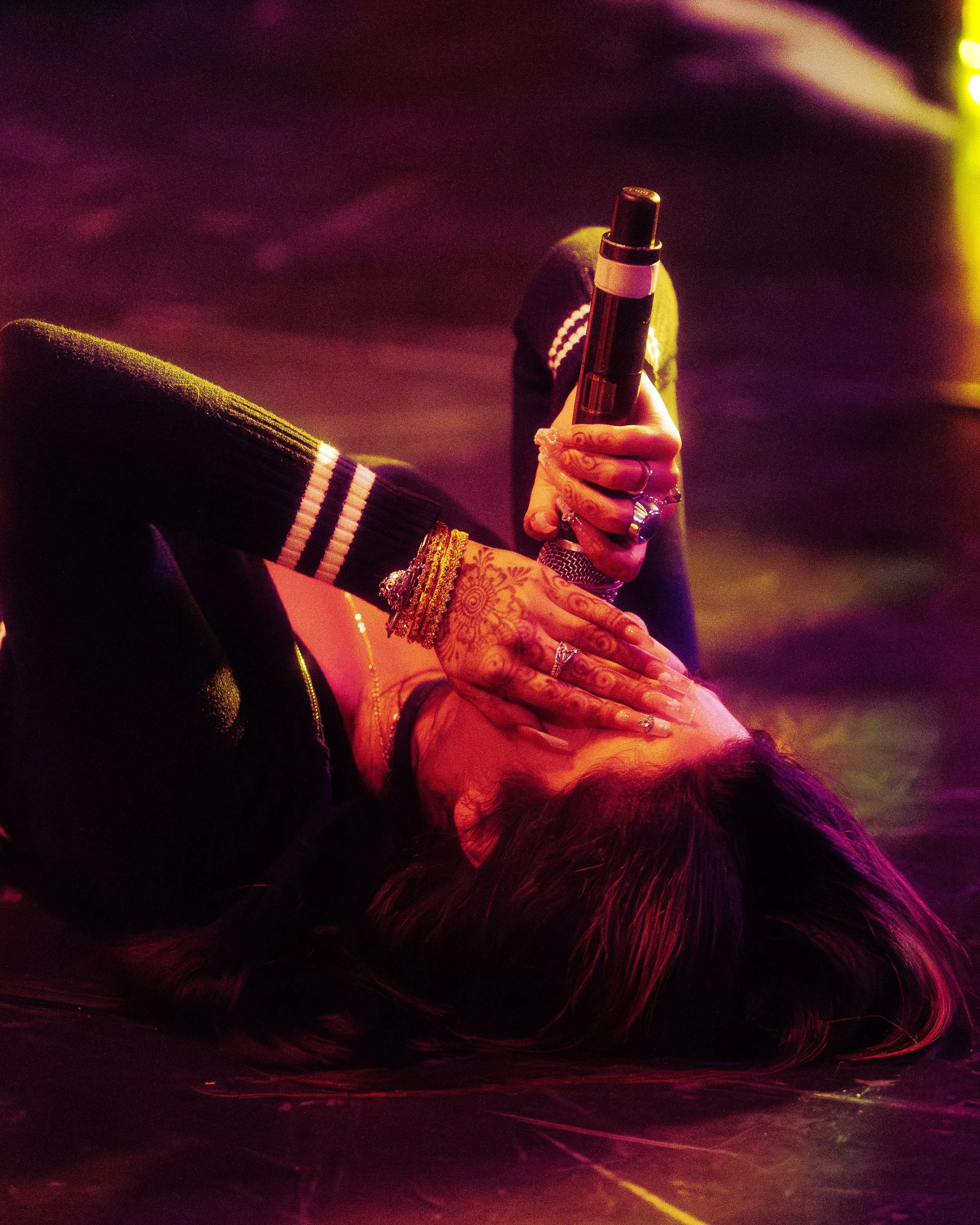 Person lying on the floor with their head turned sideways, holding a microphone above their face. They are wearing a dark jacket with striped cuffs and have colorful tattoos on their hand and wrist, along with multiple rings and bracelets. The backgr