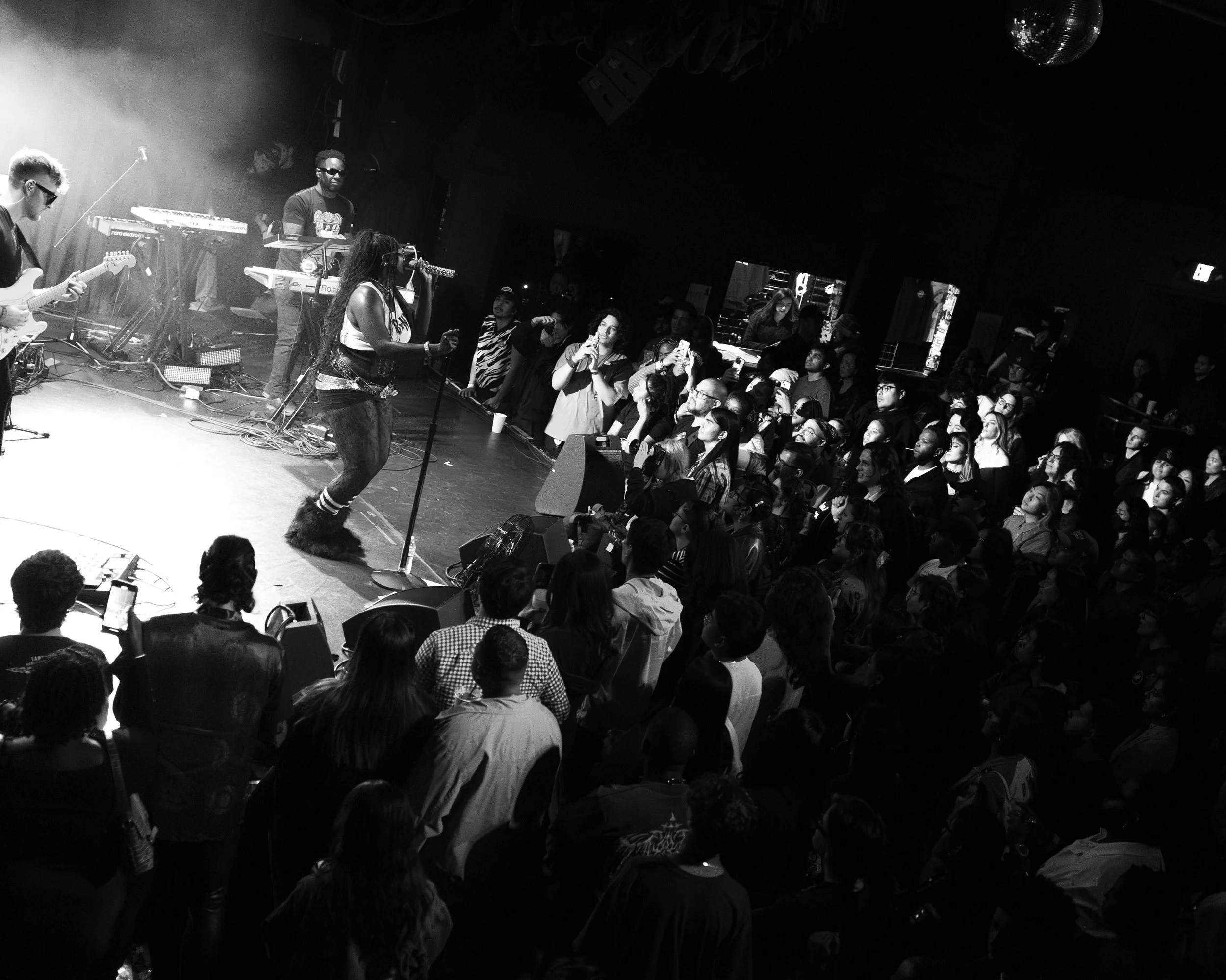 A black and white photo of a live concert with a female singer performing on stage, accompanied by guitar, keyboard, and other musicians. The audience is standing and watching the performance closely.