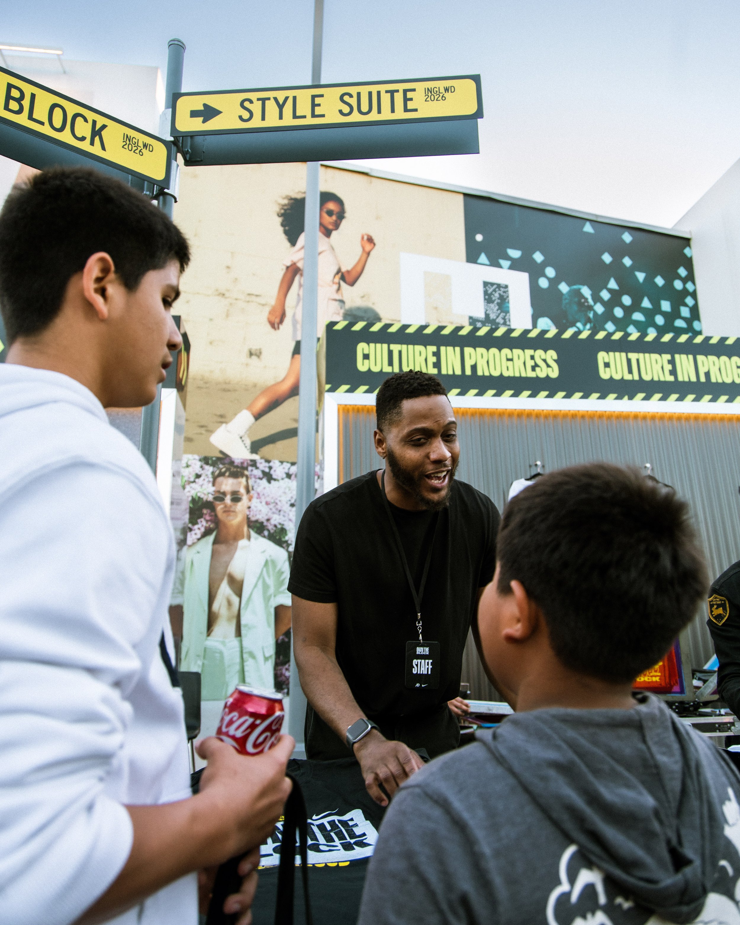 A man with a staff badge talking to two boys at an outdoor event with signages labeled "Style Suite" and "Block" and a backdrop displaying "Culture in Progress" and fashion posters.