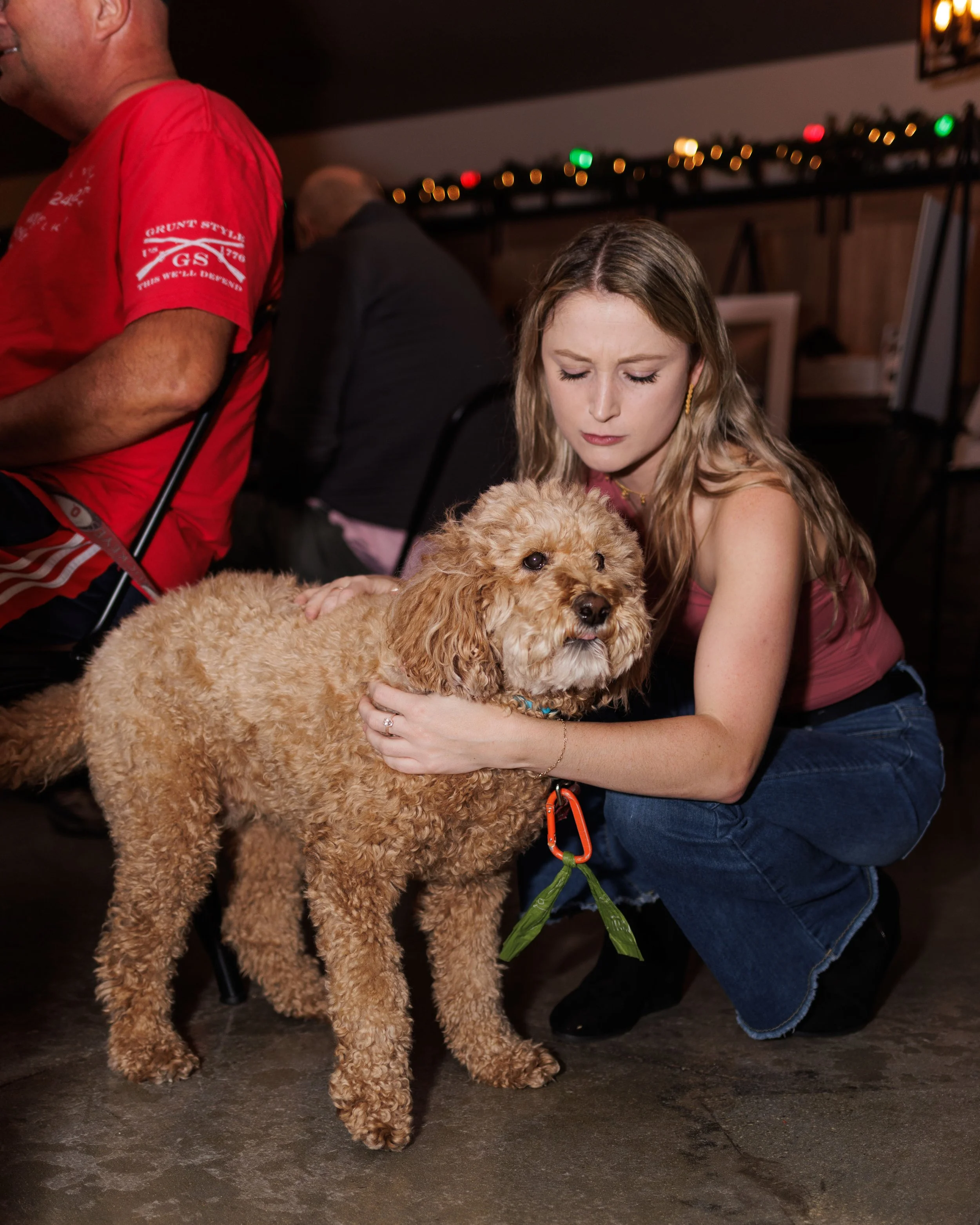 A woman crouching down, holding a curly-haired dog with a harness. The woman has long blonde hair and is wearing a pink sleeveless top and jeans. There are other people in the background, and the setting appears to be a dimly lit indoor space, possib