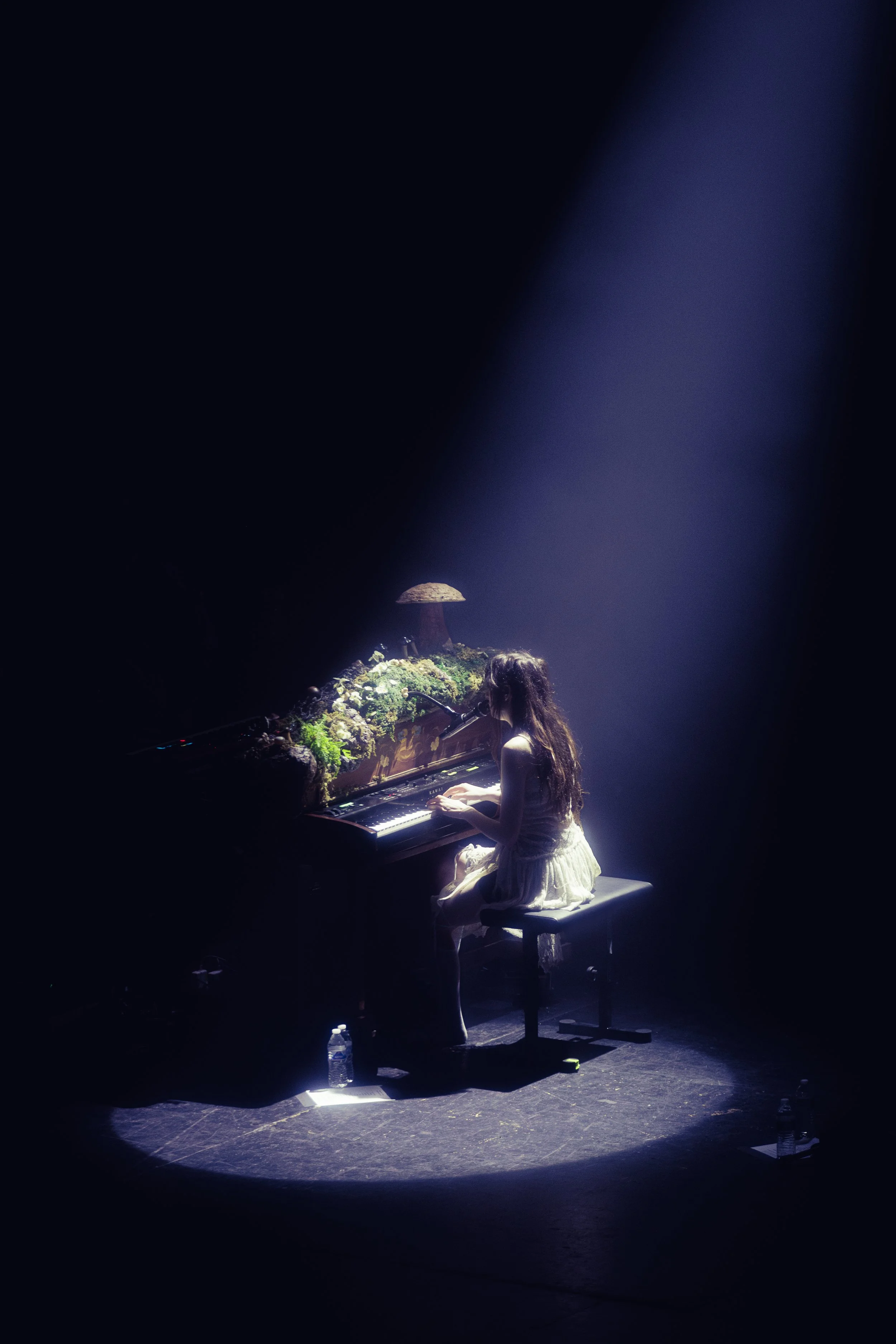 A girl in a white dress playing a piano on a dark stage, illuminated by a spotlight, with a moss and mushroom on the piano.