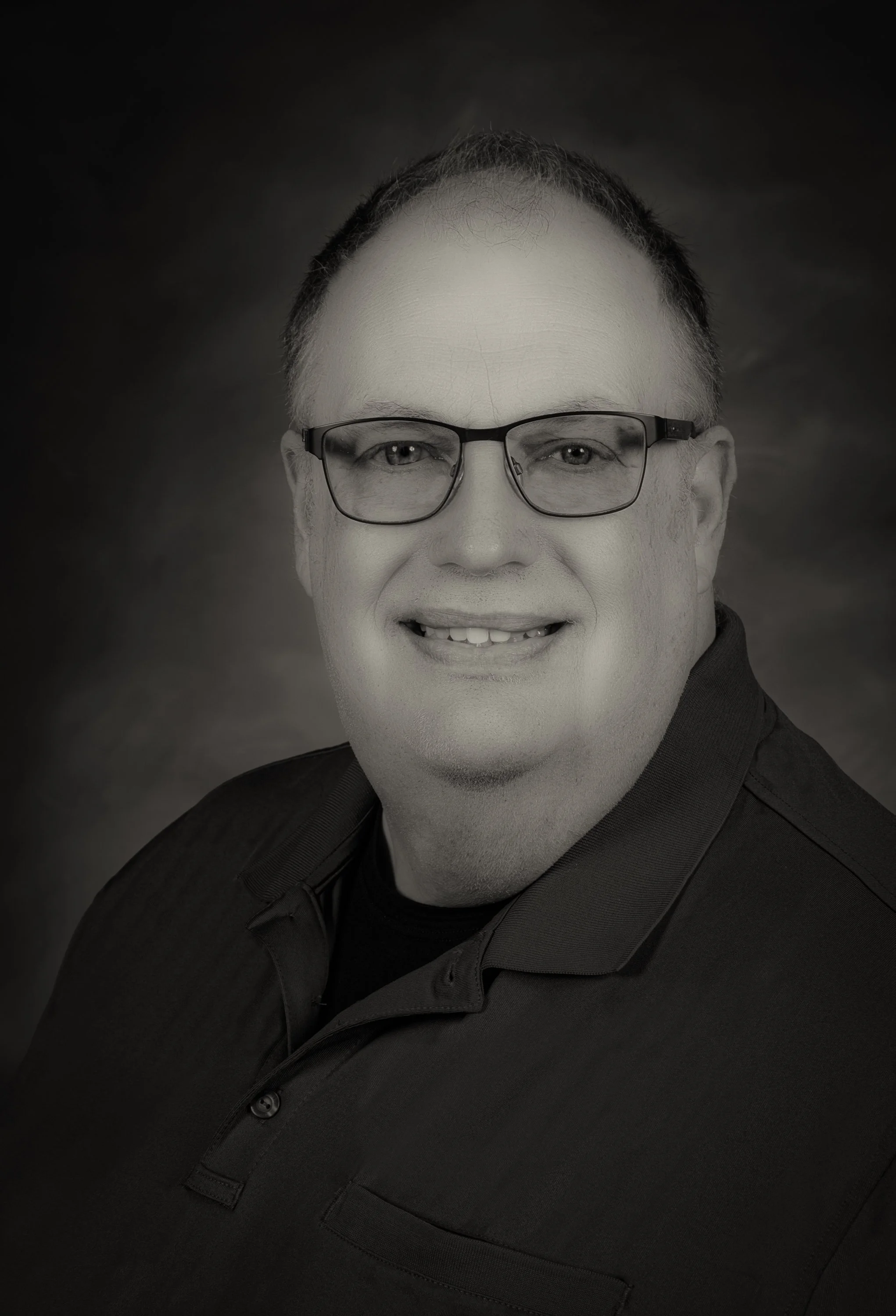 Black and white portrait of a smiling man with glasses, wearing a dark shirt, against a neutral background.
