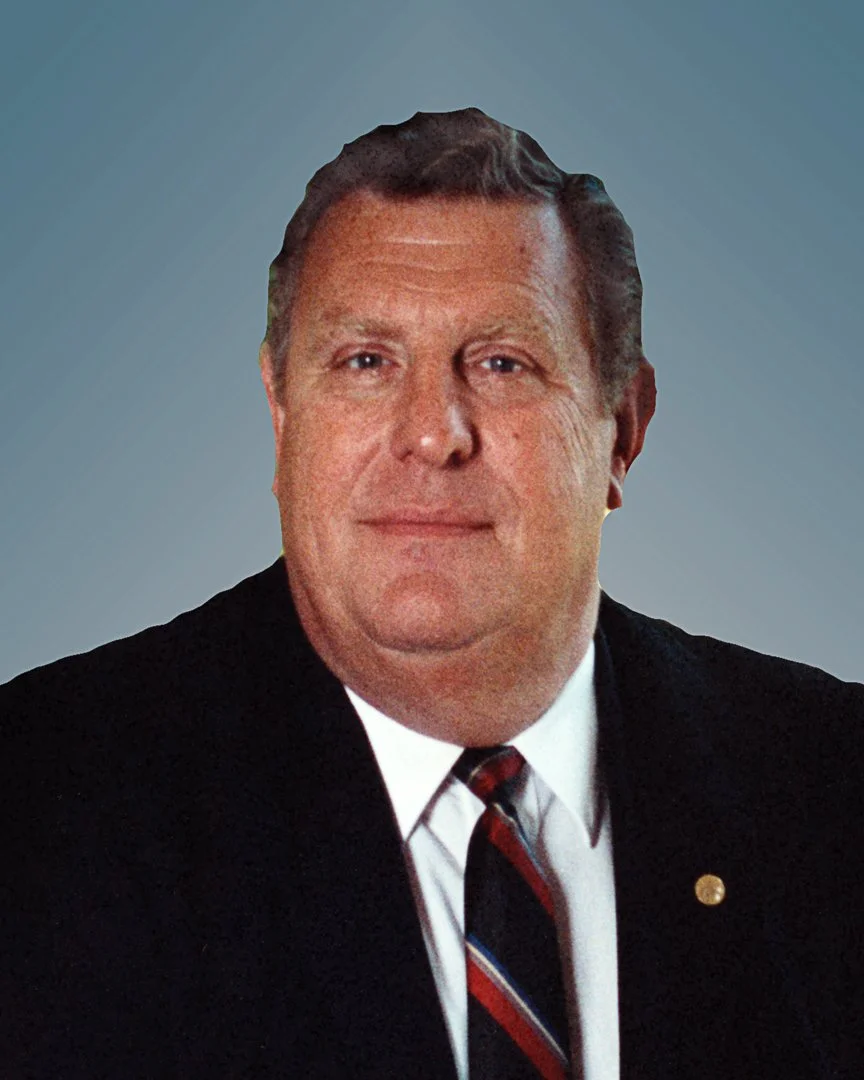 A middle-aged man with short, dark hair in a formal black suit, white shirt, and striped tie, standing against a plain gradient blue background.