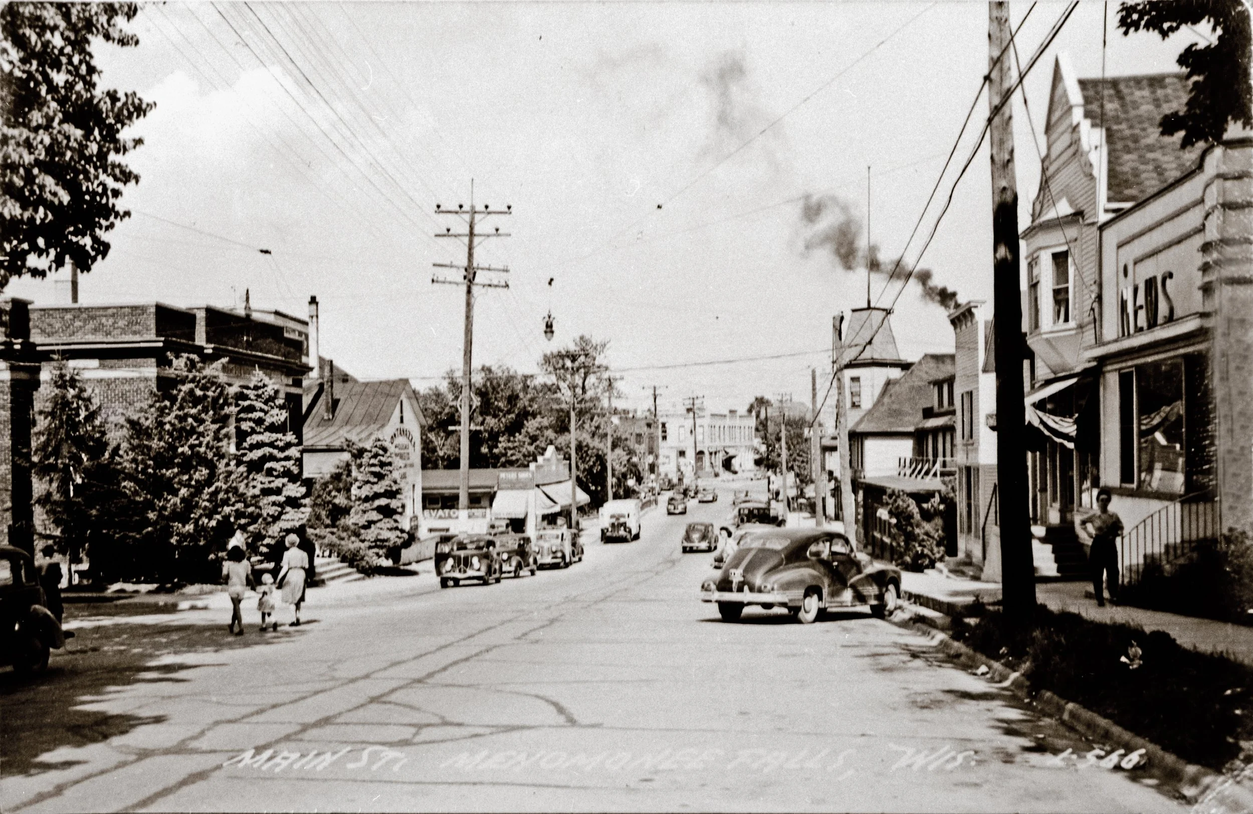 A black-and-white photo of a small town street with vintage cars parked along the sides. Several people, including children, are walking on the sidewalk. There are trees, utility poles, and buildings with signs, including a newsstand. Smoke rises from a chimney in the background. The street view is slightly inclined, showing a town scene from an earlier time period.