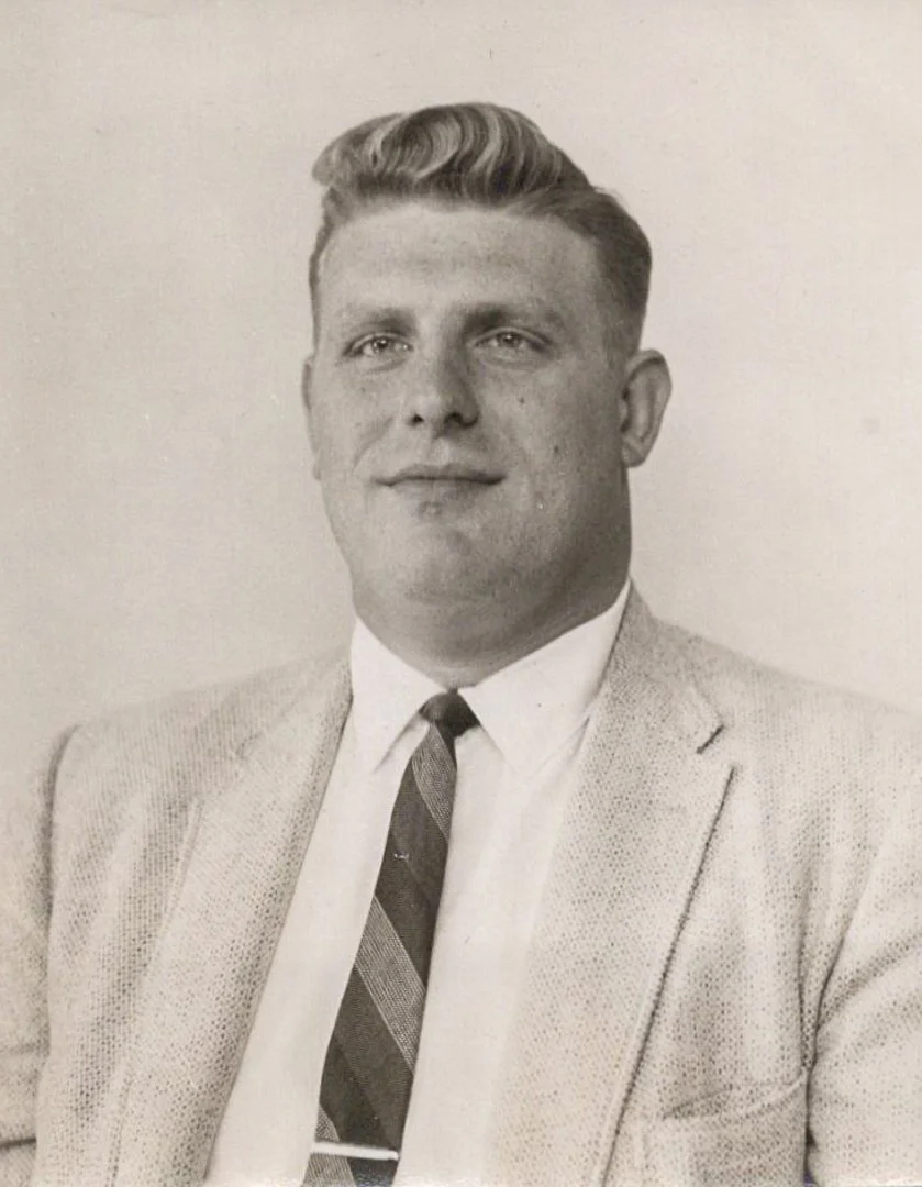 Black and white photograph of a man dressed in a suit jacket, white shirt, and a striped tie. He has neatly styled hair and is looking directly at the camera with a slight smile.