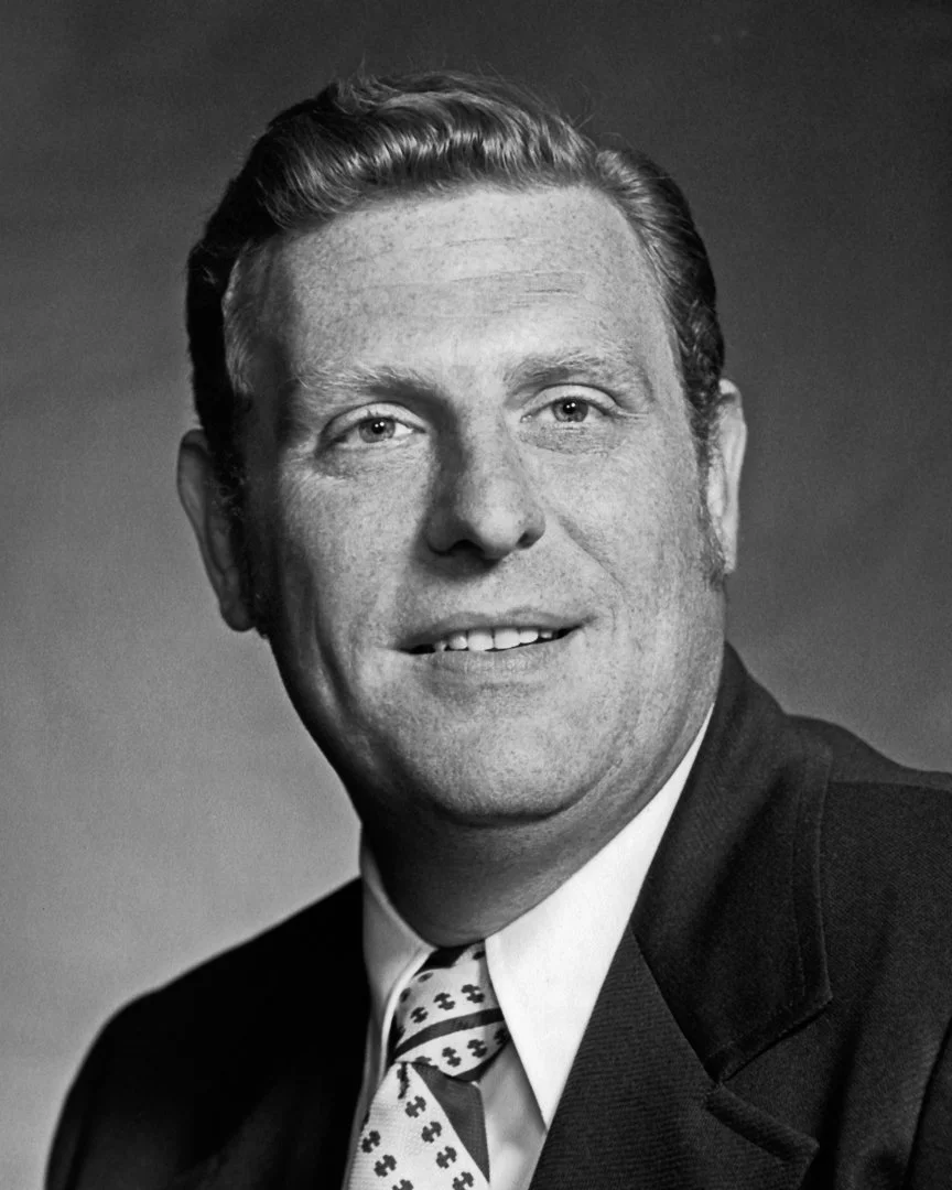 Black and white portrait of a smiling man wearing a suit and patterned tie.