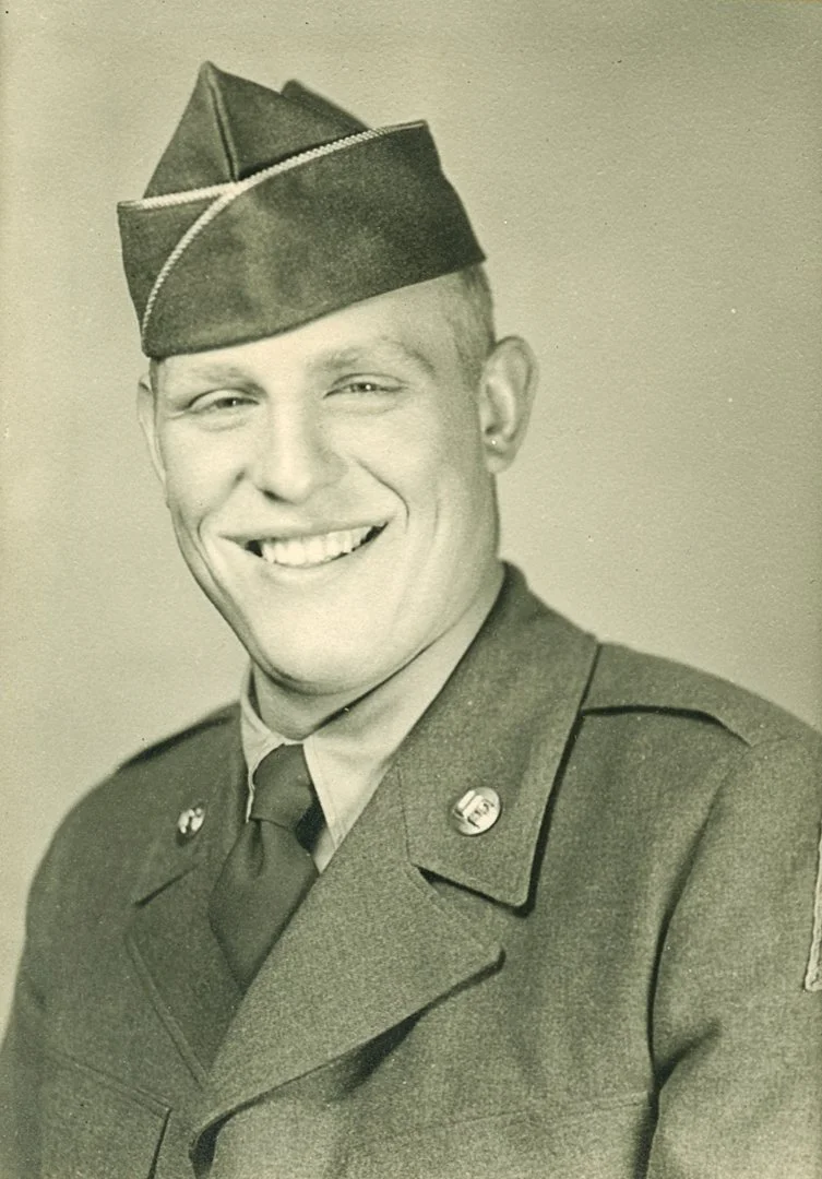 Black and white photo of a smiling young man in a military uniform with a hat, jacket, tie, and insignia.
