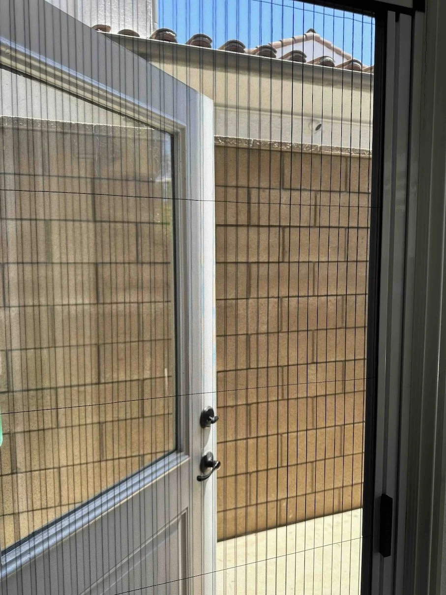 View through a screened door showing an outdoor space with a beige brick wall, a brown tiled roof, and a clear blue sky.