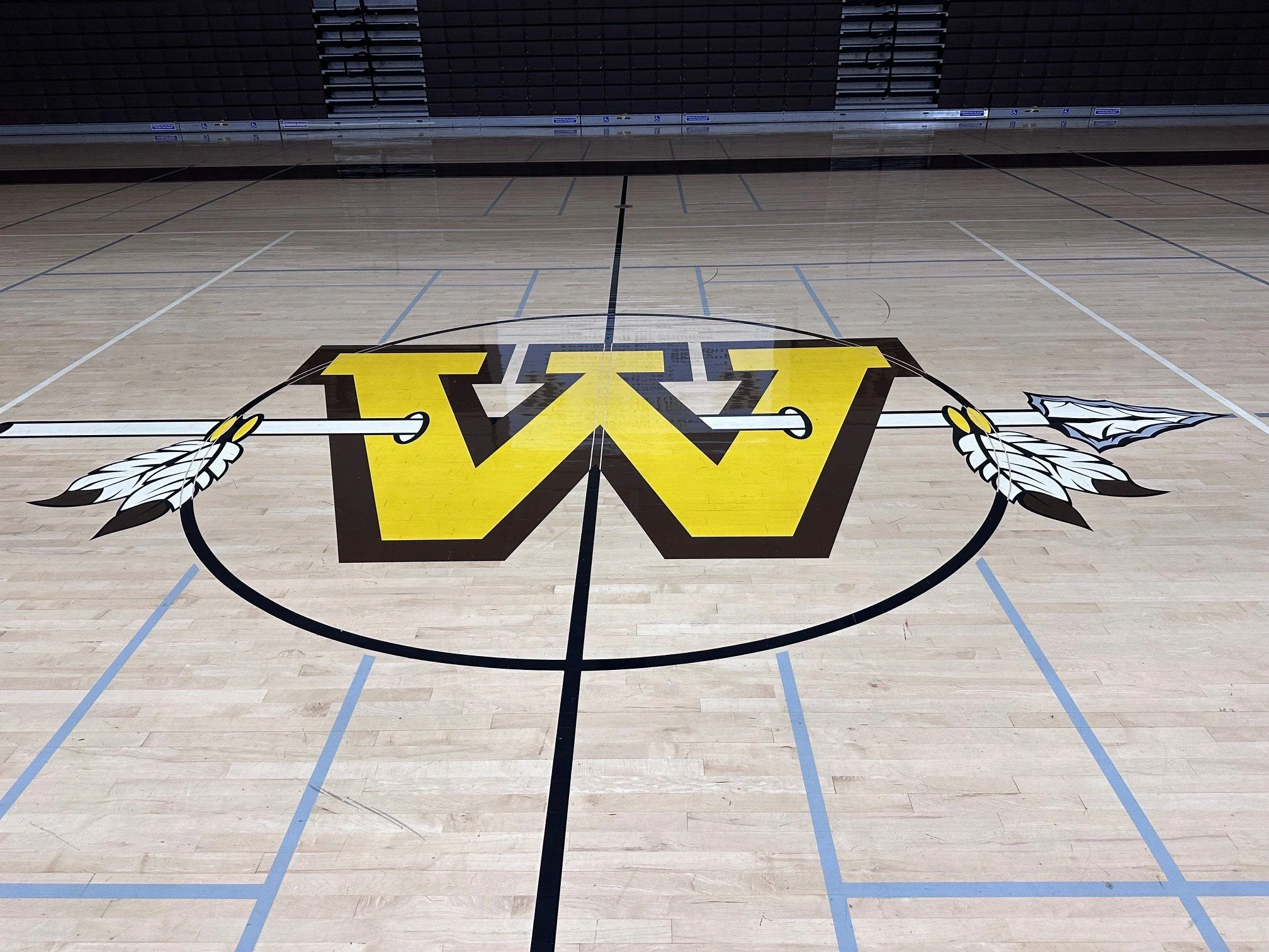 An empty basketball court with a large yellow and black logo on the floor, featuring a stylized letter 'W' and feather accents, with seating in the background.
