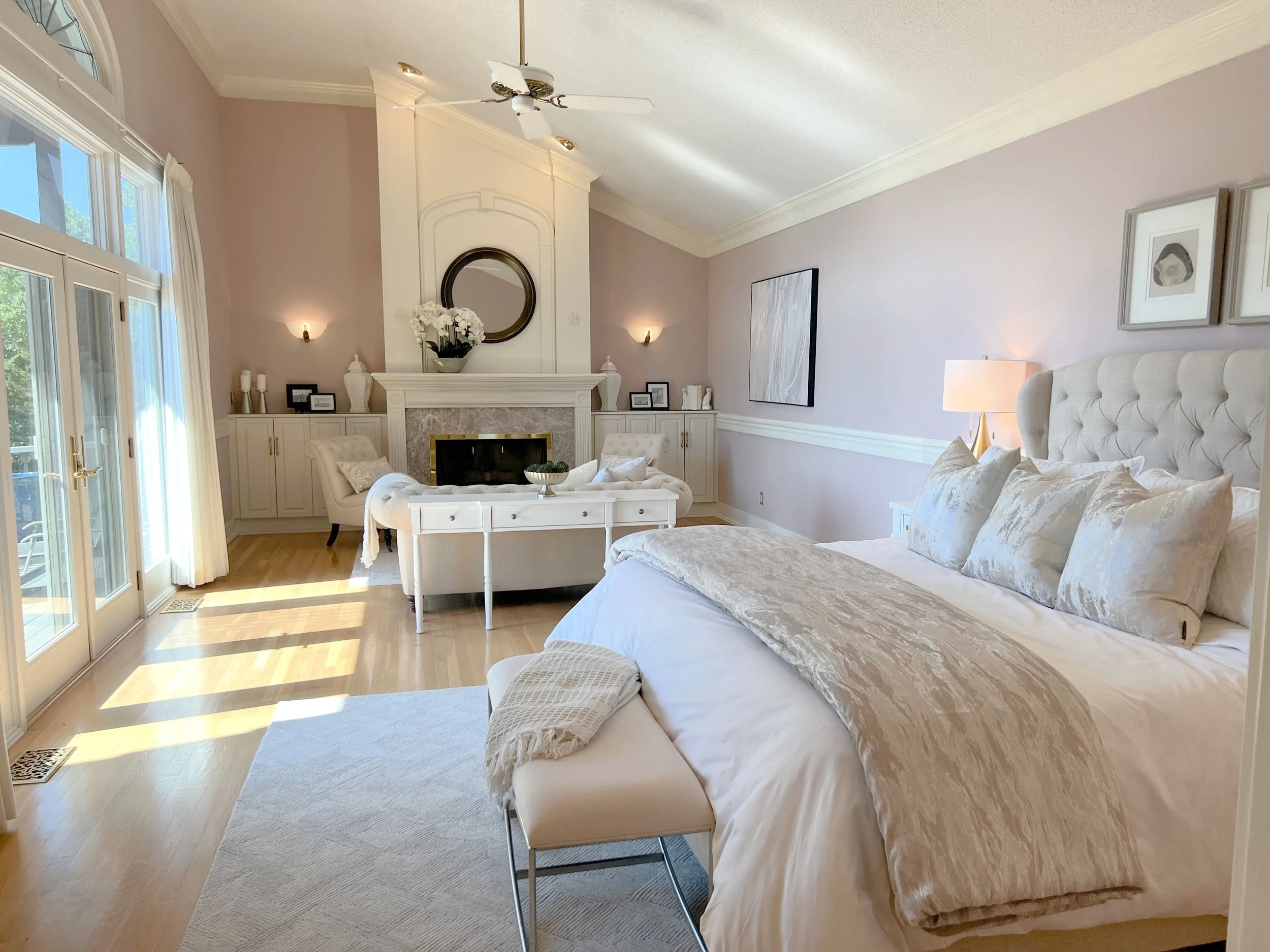 A bright bedroom with a large bed, white headboard, and plush pillows. There is a lamp on a nightstand and artwork on the walls. A sitting area with a fireplace, mirror, and decorative items is visible across the room. Sunlight streams through glass 