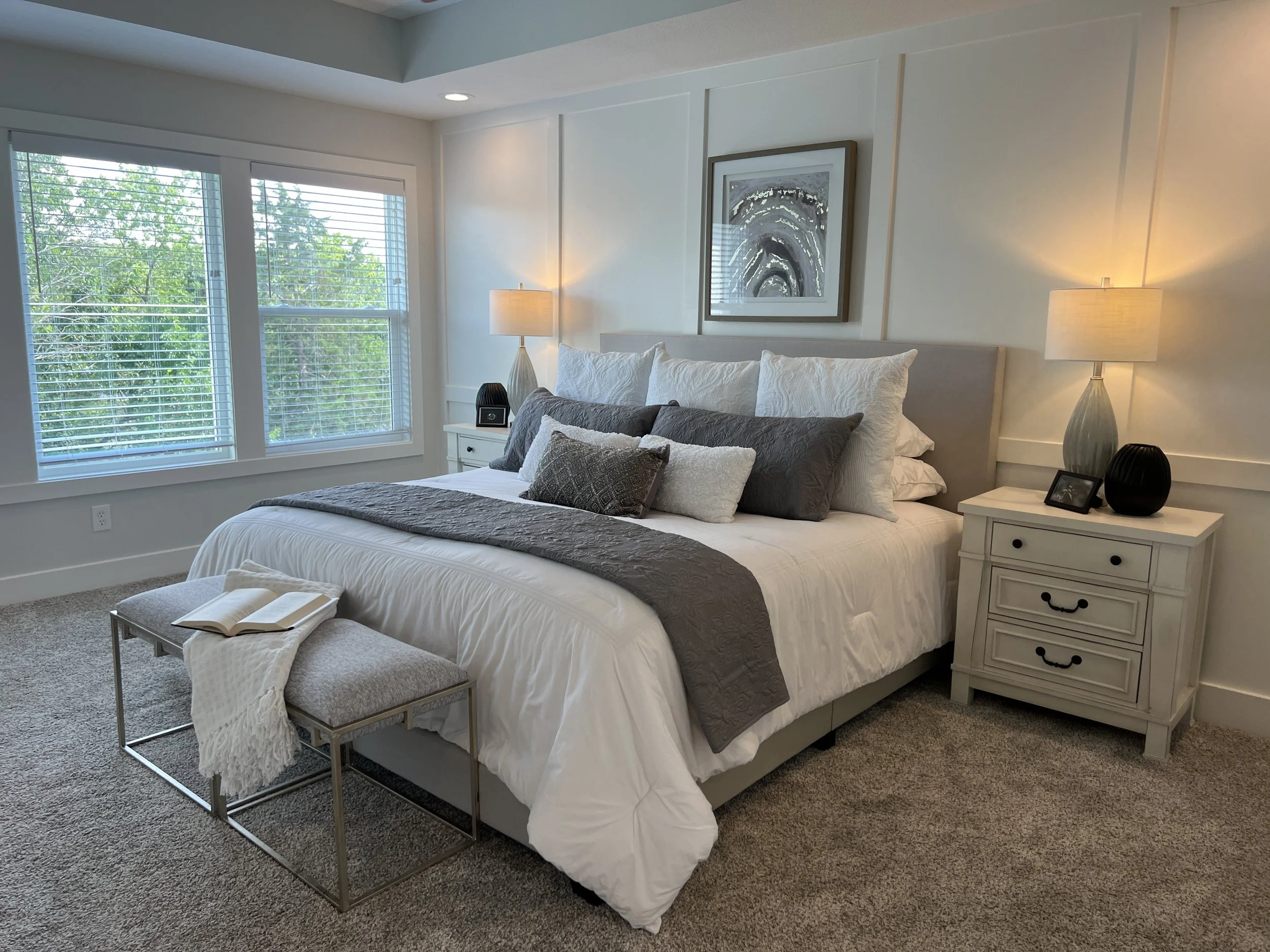 A neatly made bed with white and gray bedding, decorative pillows, and a bench at the foot of the bed with a book and throw blanket. Two white nightstands with lamps flank the bed, and large windows with blinds offer a view of green trees outside.