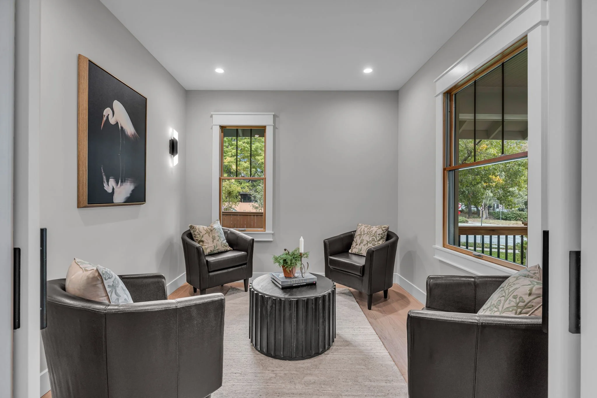 A small, modern sitting room with four black leather armchairs with decorative cushions, a round black coffee table with a plant and books, two large windows showing trees outside, and a piece of artwork with a stork on the wall.