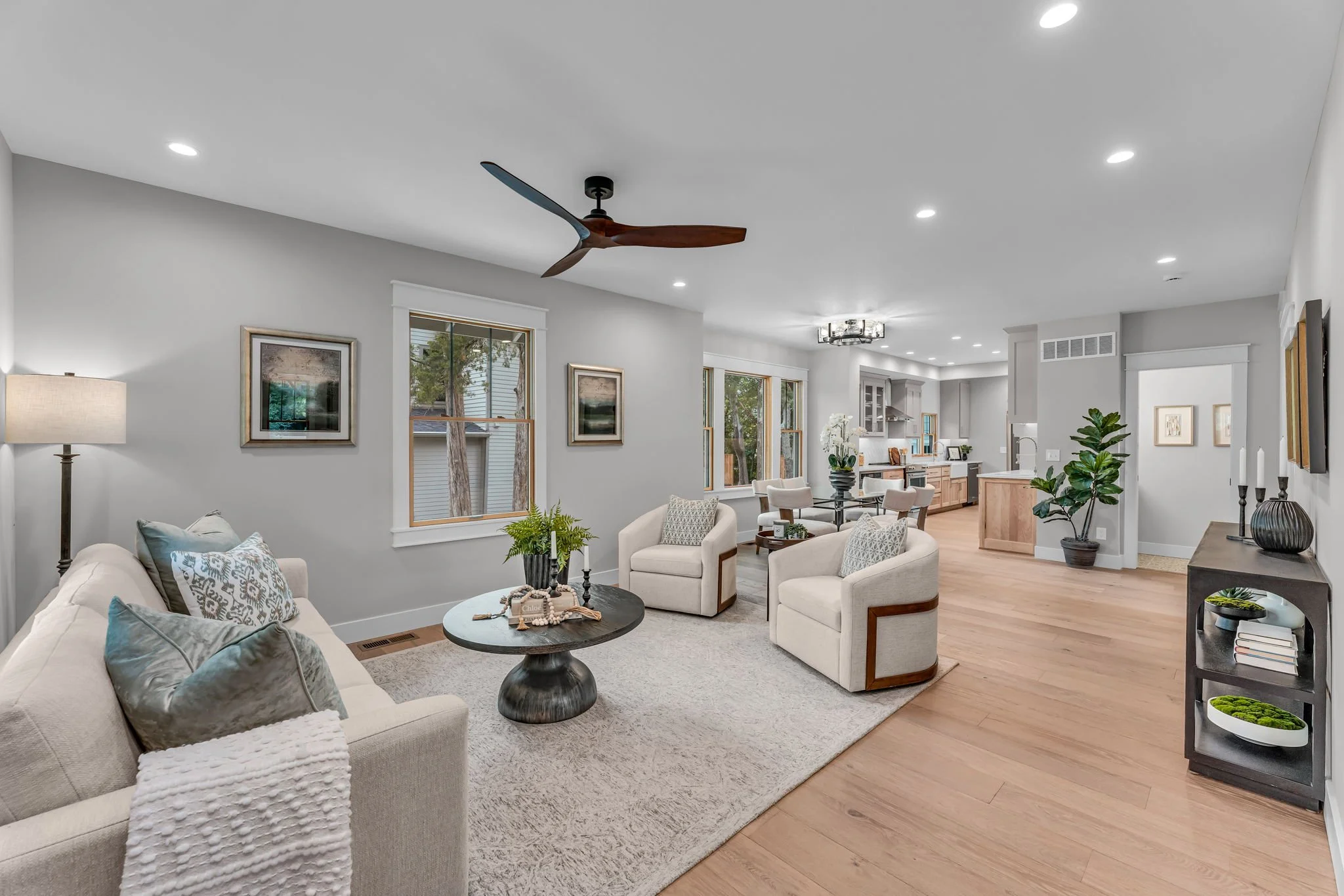 Open-concept living room with white sofa and armchairs, coffee table, ceiling fan, large windows, and visible kitchen area.