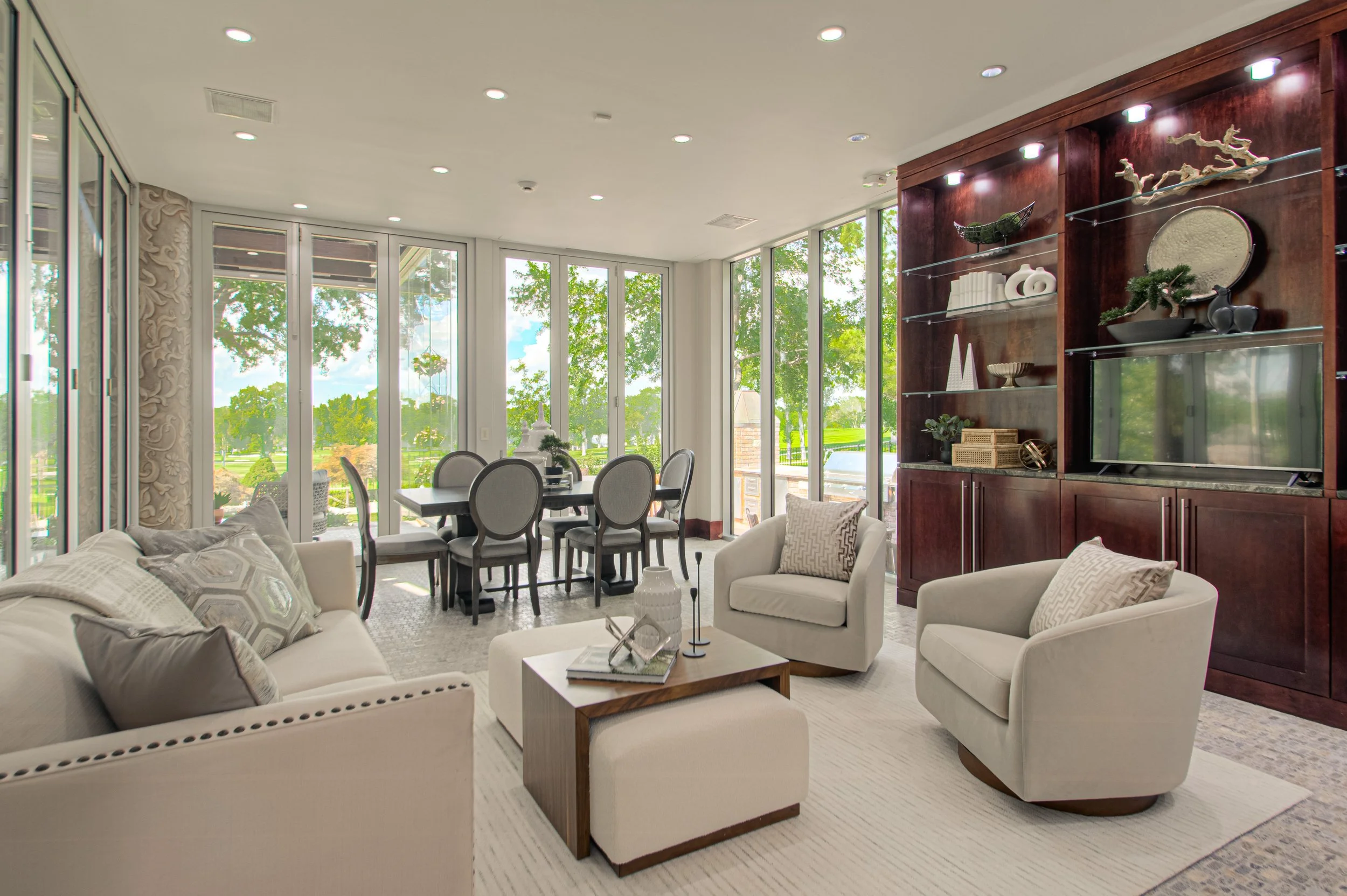 Living room with white sofa and armchairs, wooden coffee table, built-in wooden shelves with decorative items, large glass windows overlooking a green outdoor area.