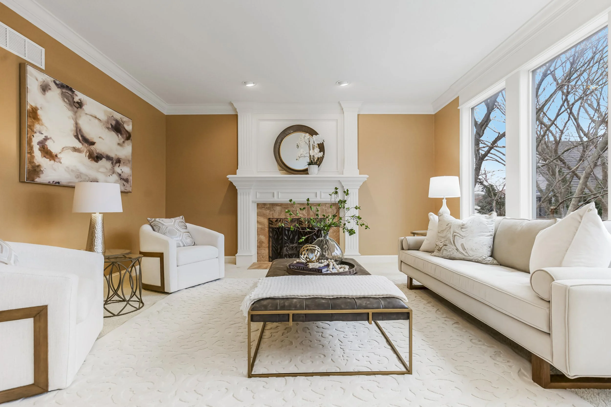 A living room with beige walls, three white sofas, a fireplace with a mirror above, large windows with a view of trees, and a coffee table with decorative objects.