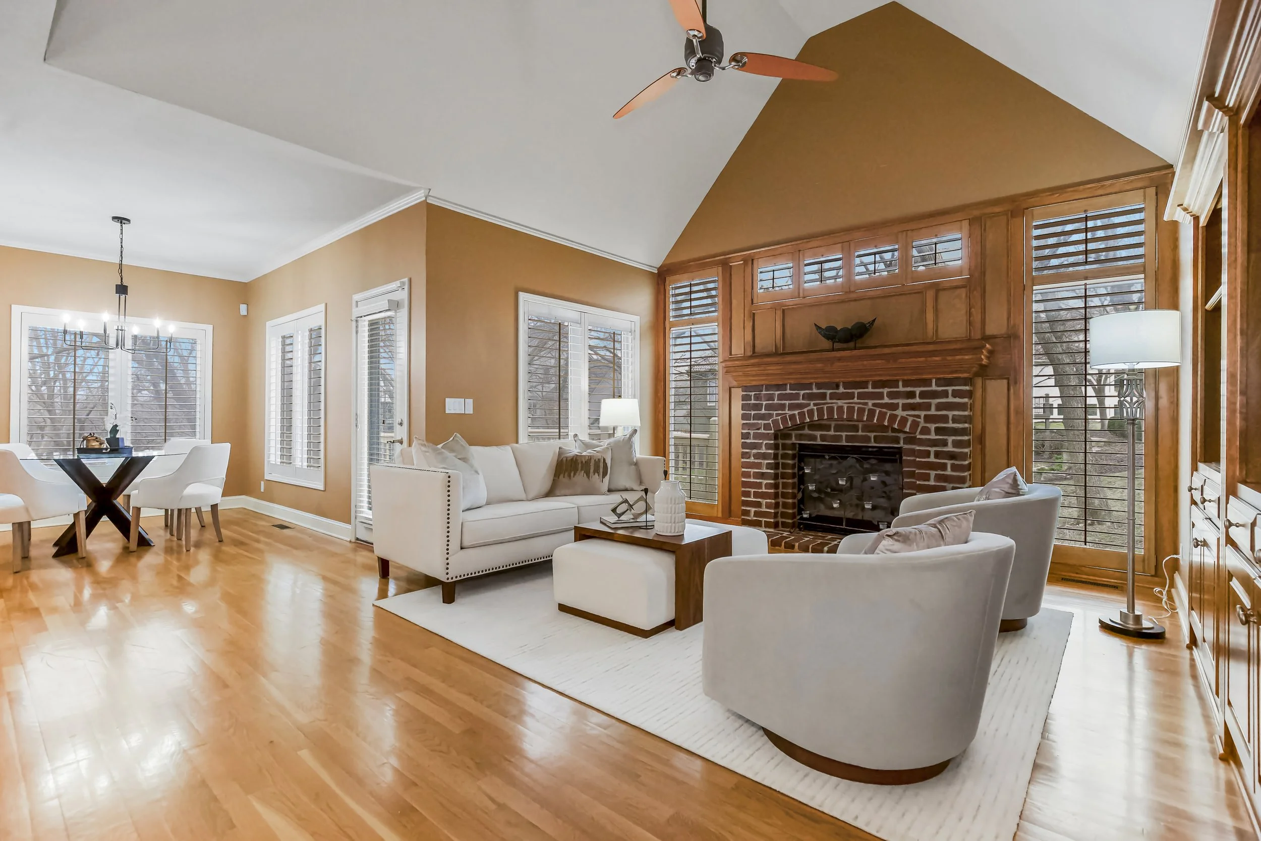 Living room with a brick fireplace, white sofa and armchairs, hardwood floors, large windows with blinds, a ceiling fan, a floor lamp, and a dining area with a chandelier.